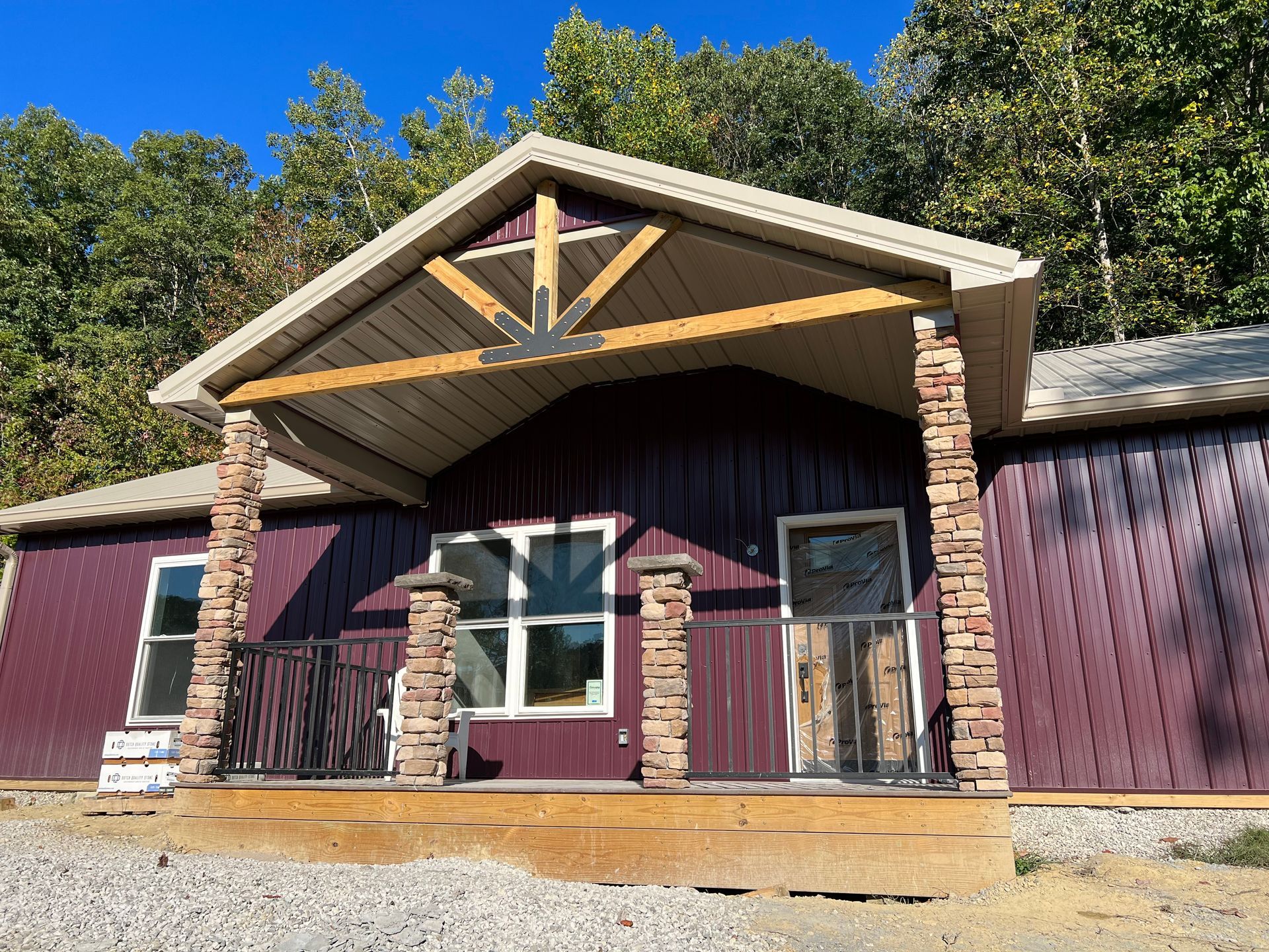A maroon cabin with a porch, stone columns, and a wooden beam above the door. The porch has a black railing and the building is surrounded by trees.