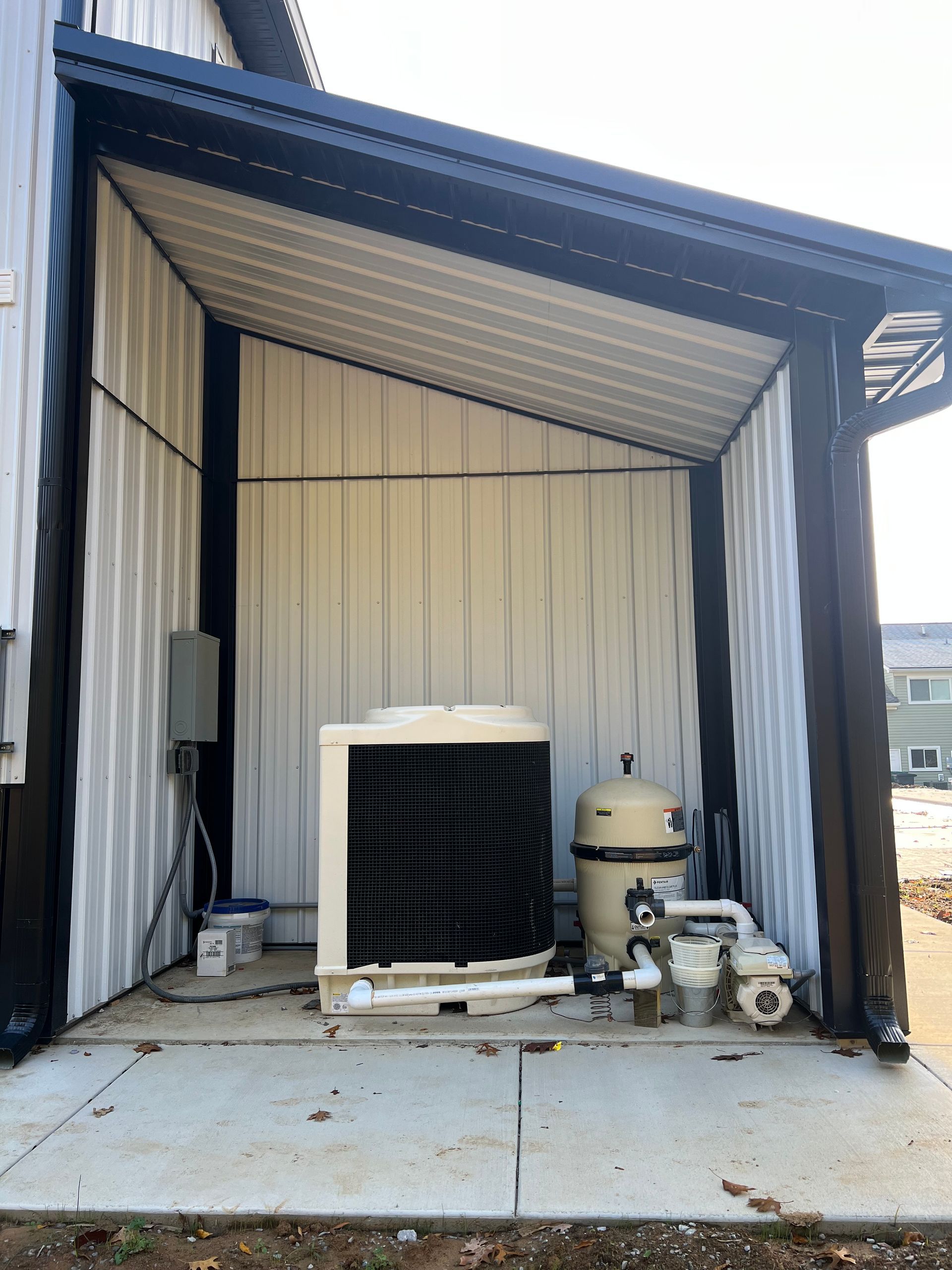 Pool equipment, including a heater and filter, housed under a white and black metal awning against a building.