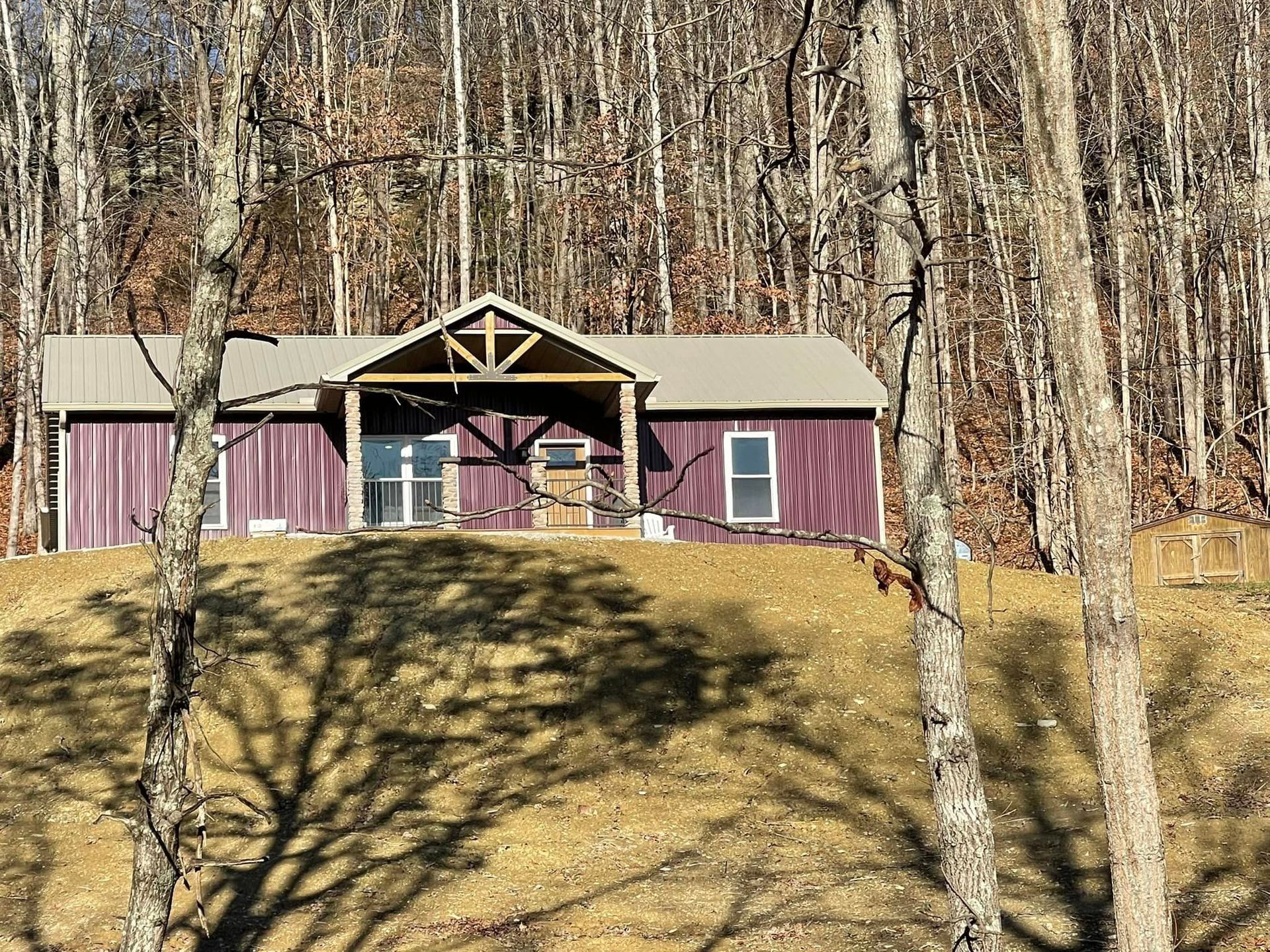 A cabin with a burgundy exterior sits on a hillside among bare trees. A wooden porch shelters the entrance.