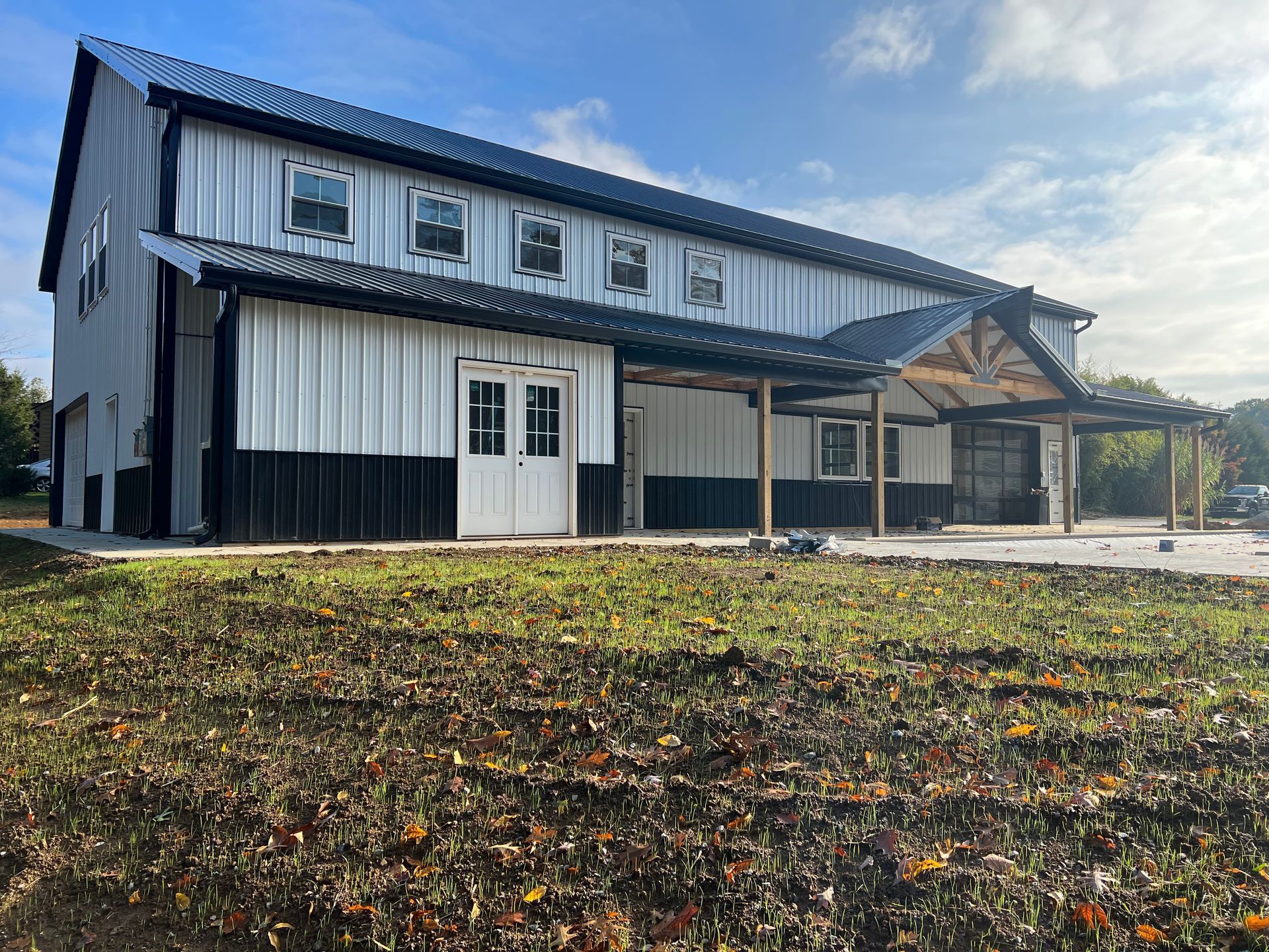 A two-story, white metal building with black accents. It has a covered porch with wooden supports on the right and sits on a grassy area.