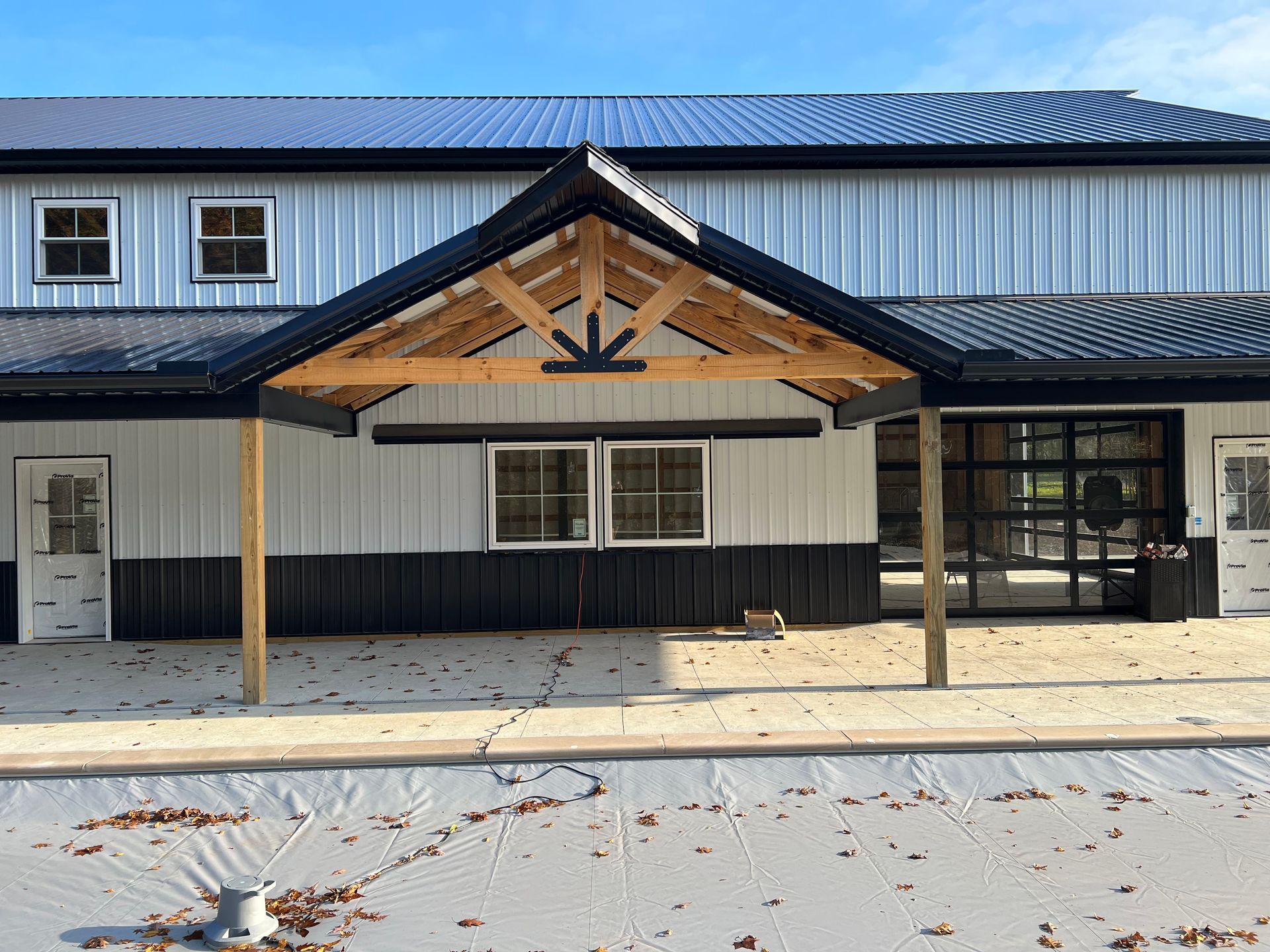 A modern white and black metal barn with a covered porch featuring a wooden truss, concrete patio, and large windows.