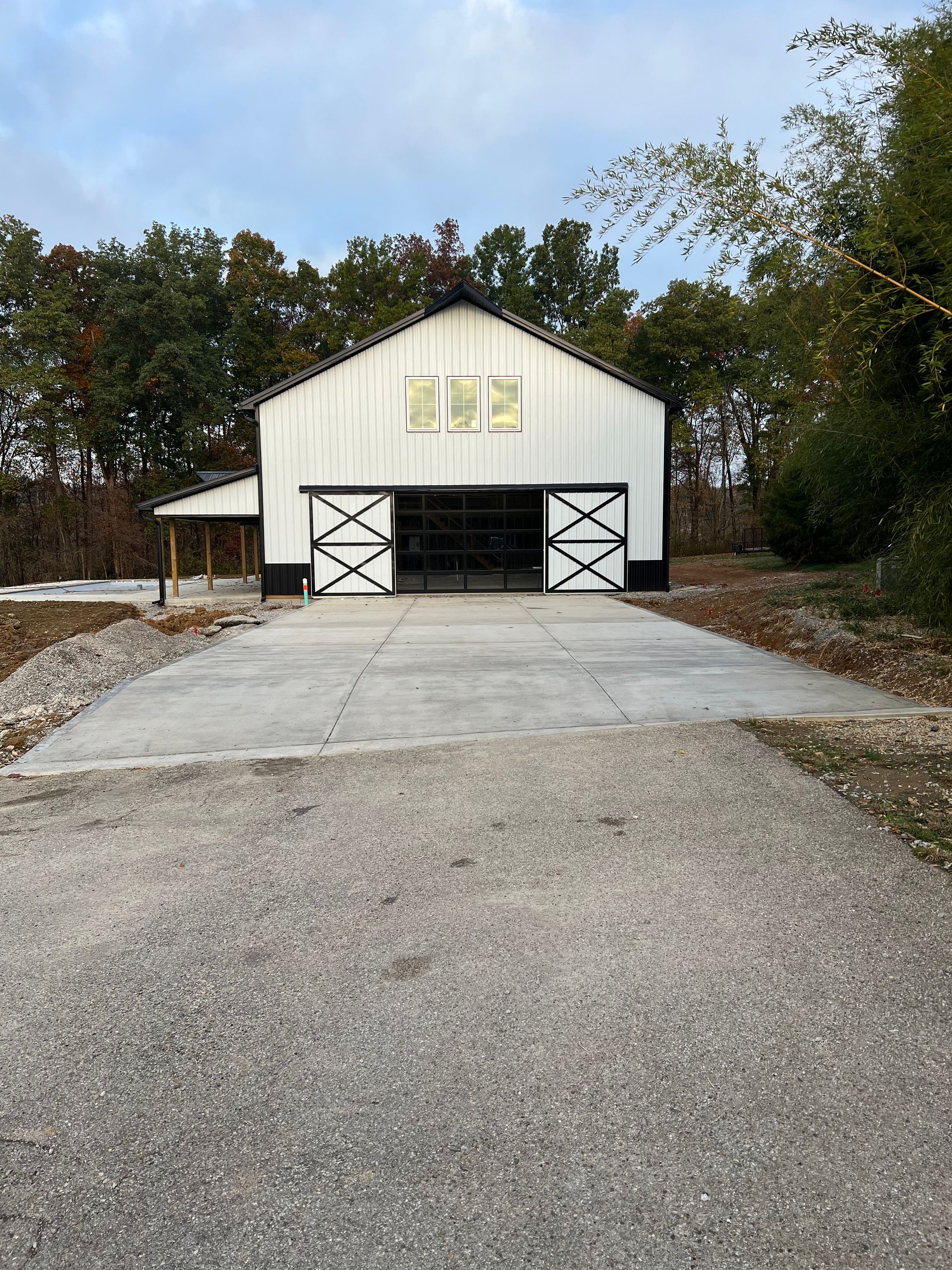 White barn with black trim, open doors, and concrete driveway. Gravel road leads up to the barn in a wooded setting.