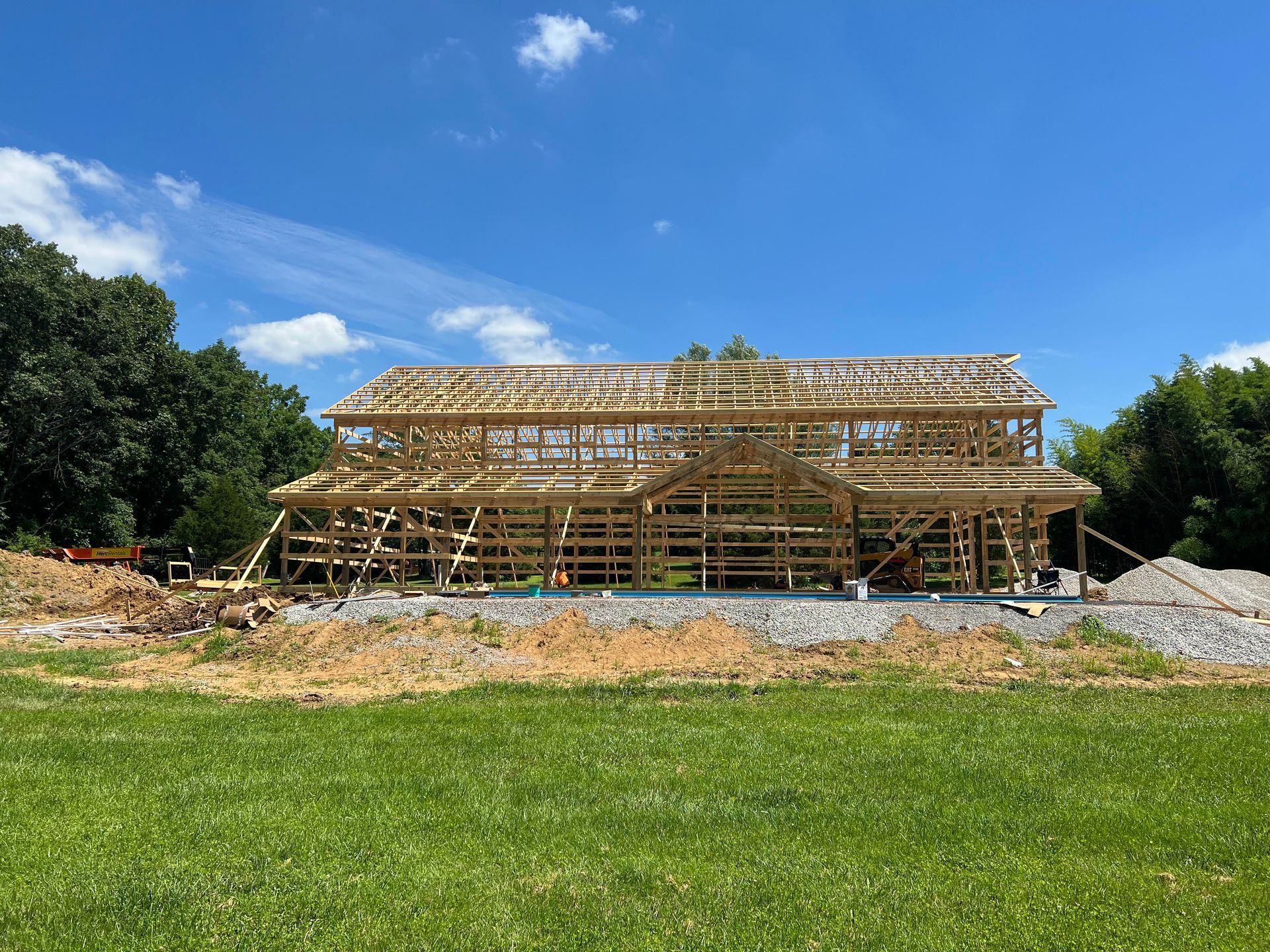 A two-story house under construction, framed with wooden beams against a bright blue sky. The house sits on a gravel base, surrounded by grass and trees.