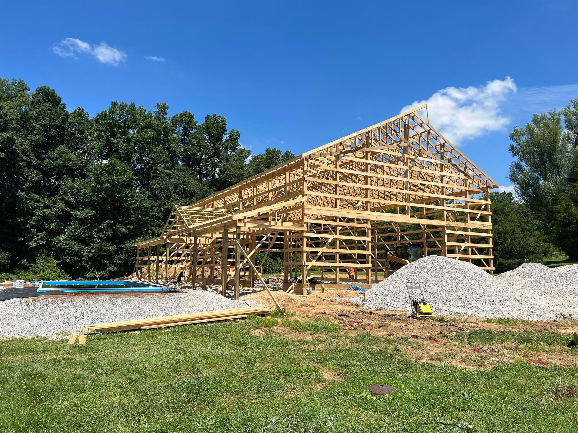 Wooden barn frame under construction against a blue sky, with piles of gravel and trees in the background.