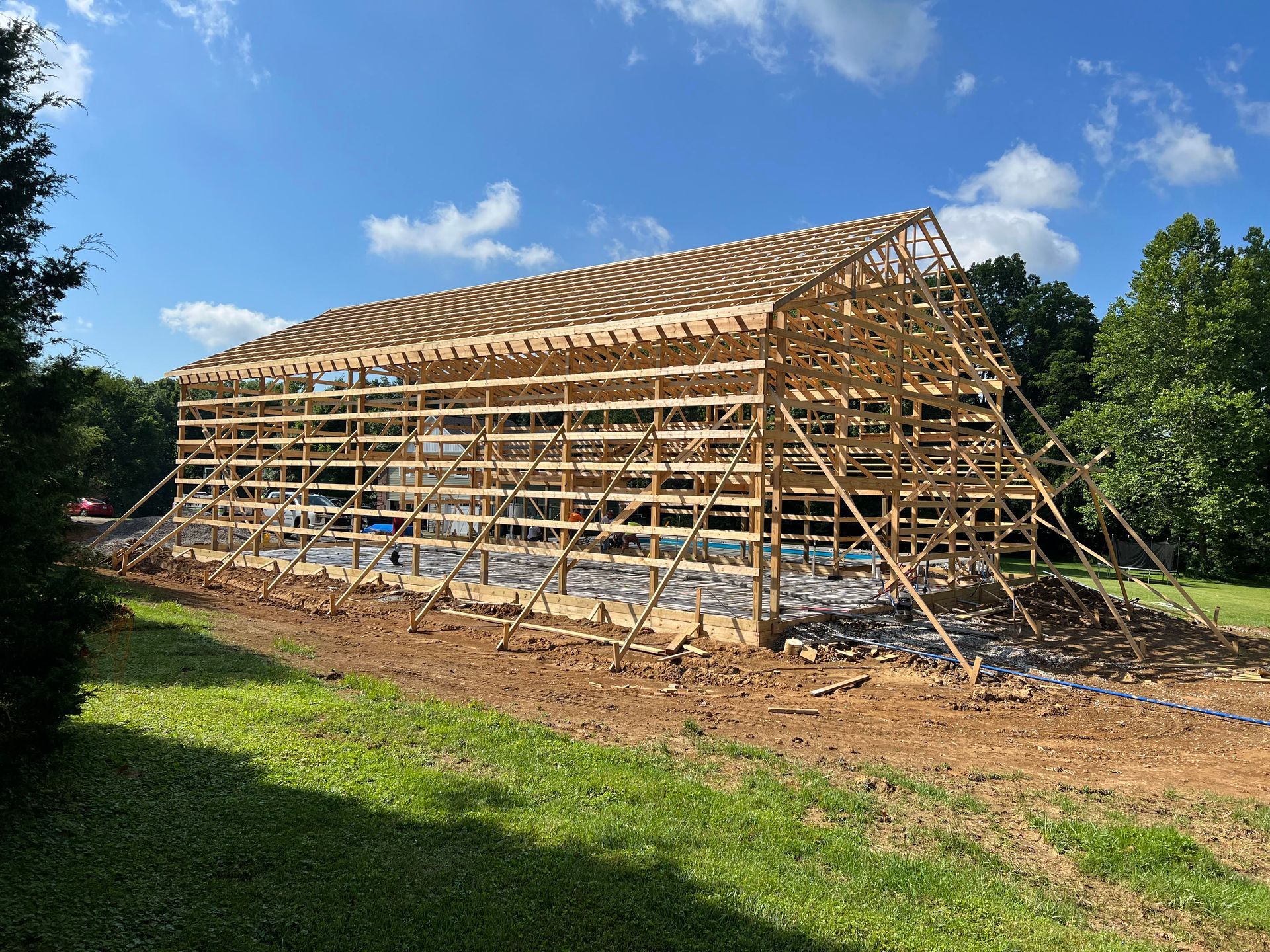 A wooden barn under construction on a grassy lot against a bright blue sky. The frame is nearly complete, with arched roof beams visible.