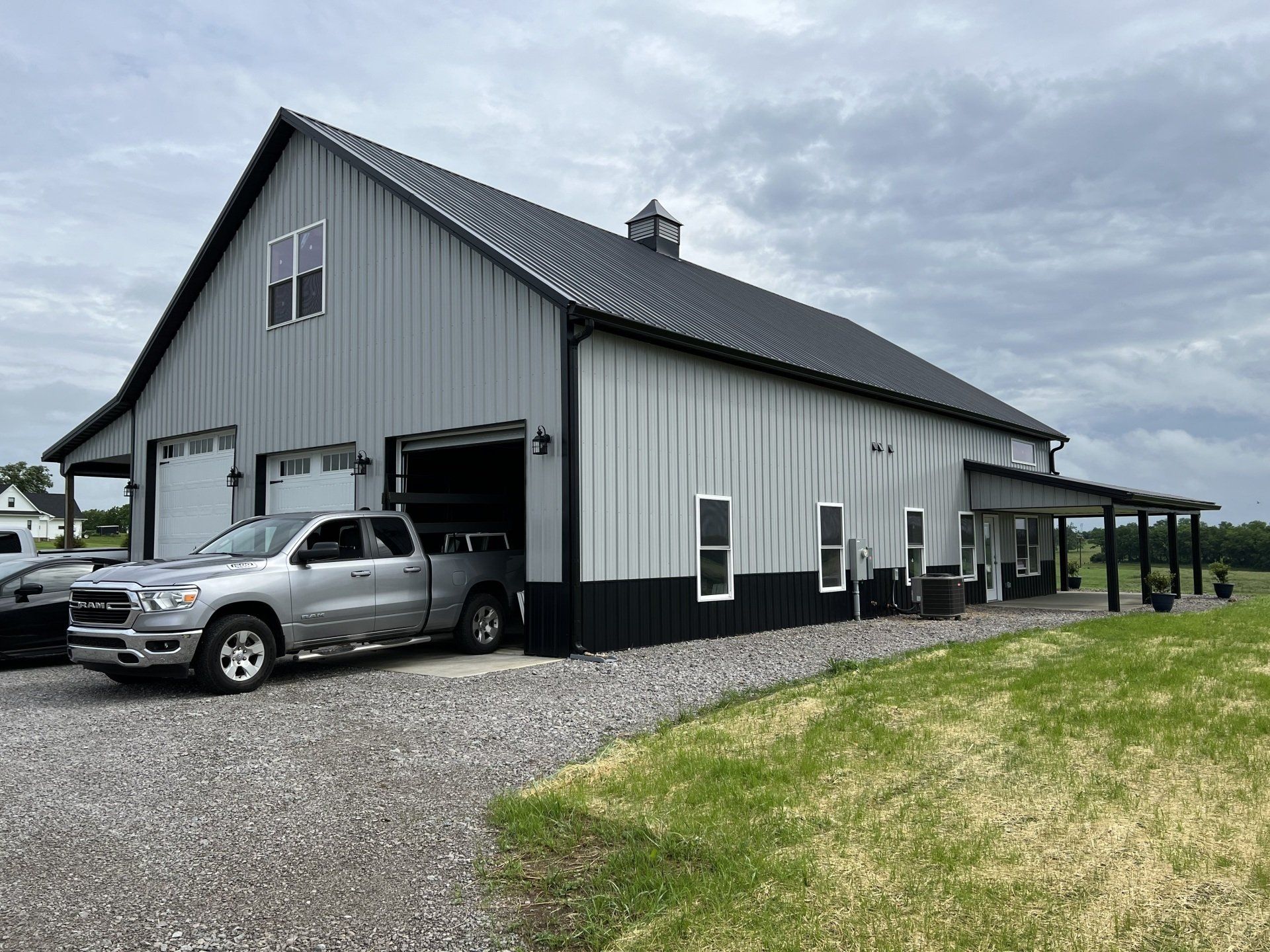Gray metal barn with black trim and a pickup truck parked in a garage. Includes a porch and green lawn.
