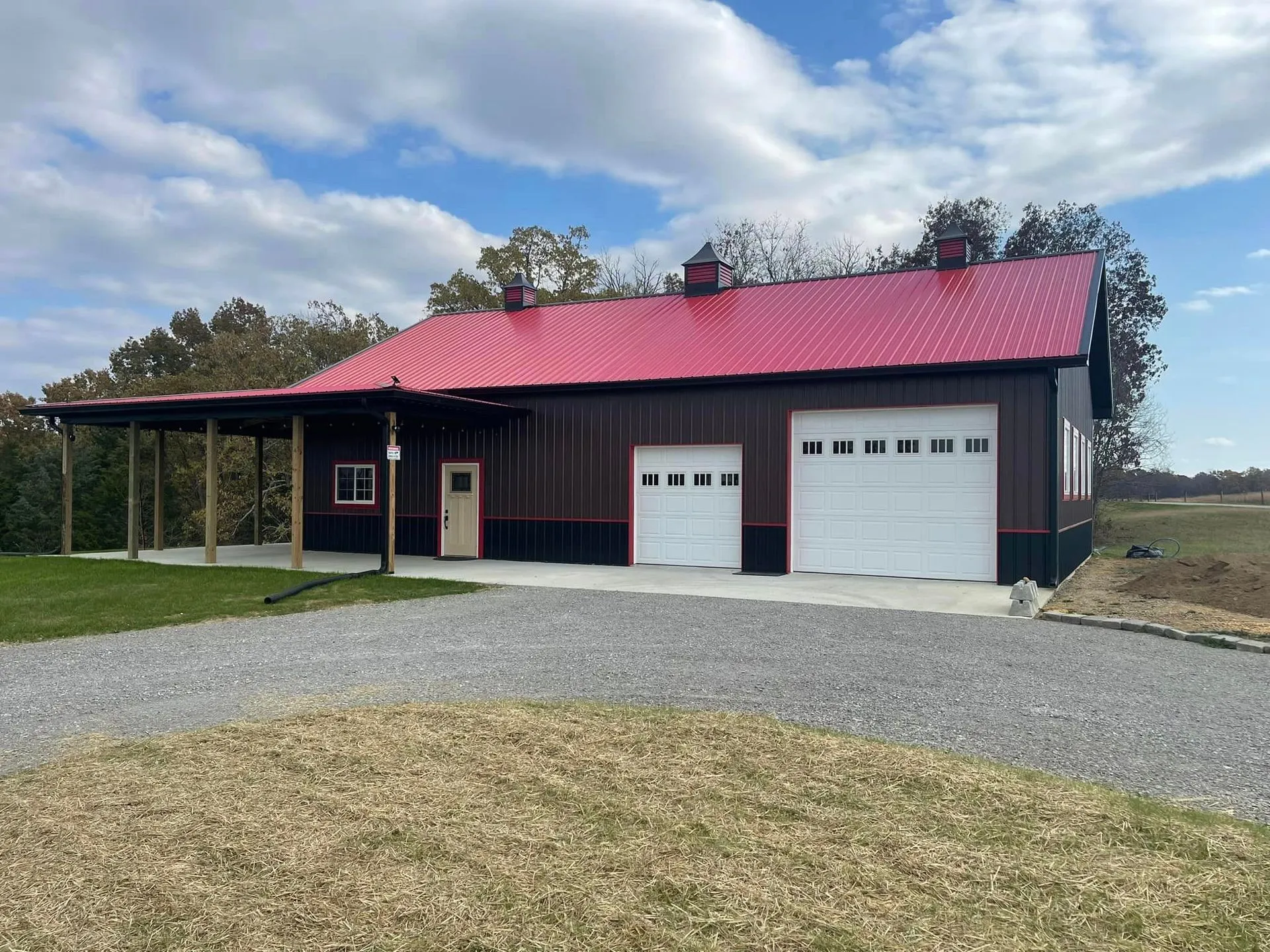 A dark brown metal building with a red roof, two white garage doors, and a covered porch, set in a grassy area under a cloudy sky.