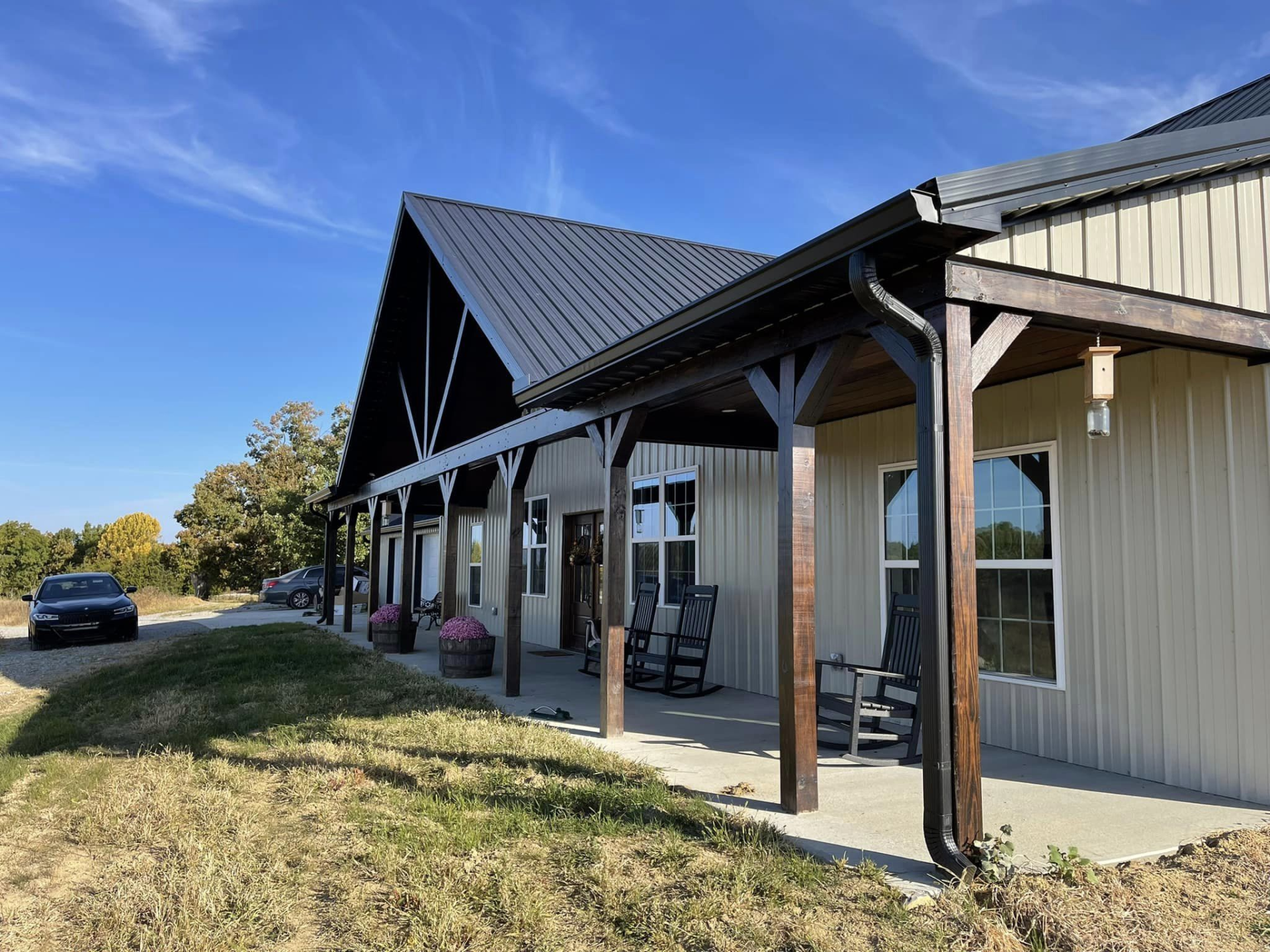 A light beige building with a black metal roof and dark brown trim. A porch has rocking chairs and faces a gravel driveway on a sunny day.