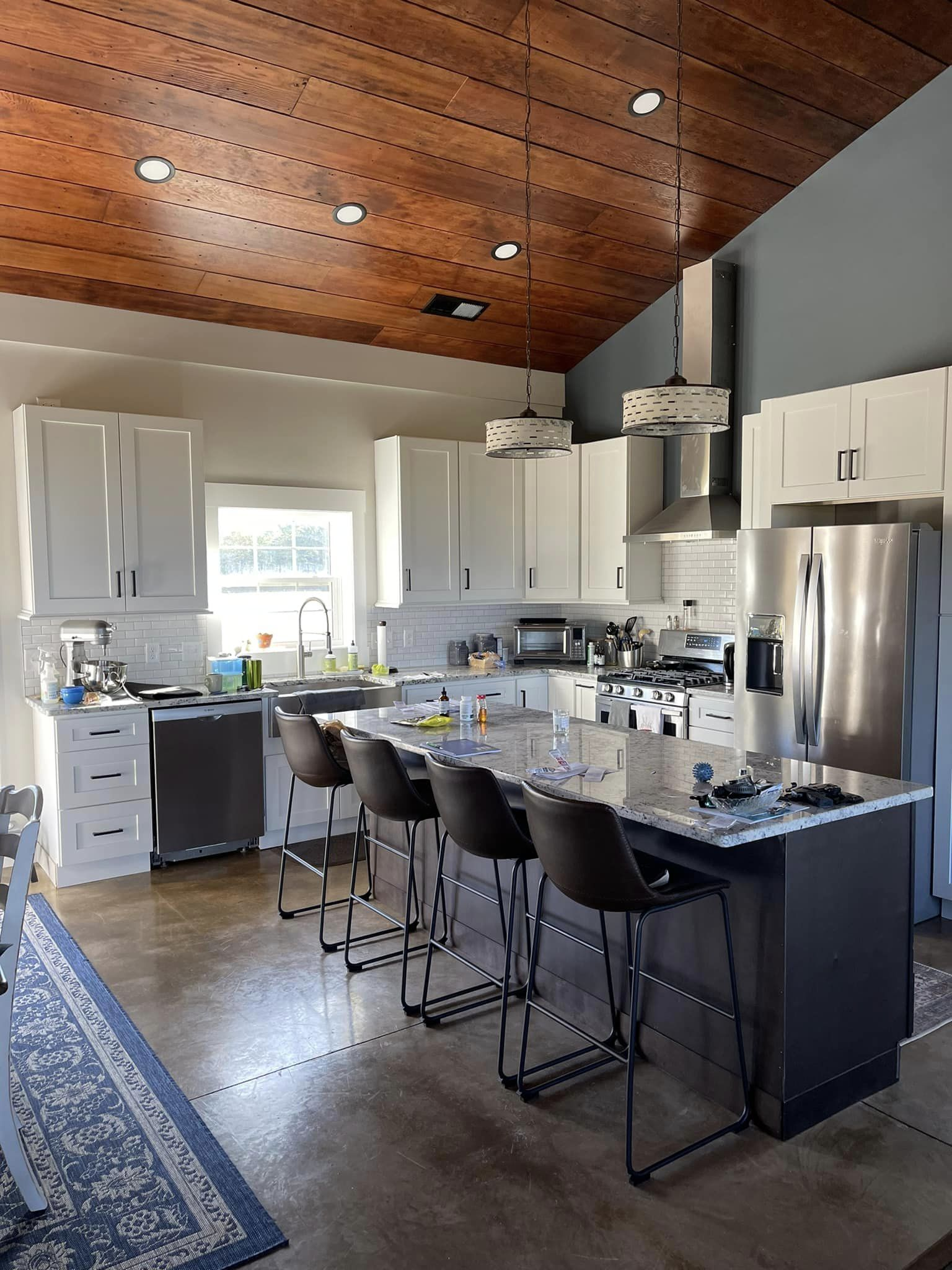 Modern kitchen with white cabinets, dark island, and stainless steel appliances. A wooden ceiling and concrete floor are visible.