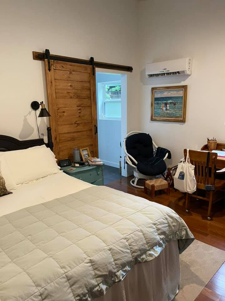 Bedroom with a bed, barn door, chair, desk, and artwork. Light gray bedding, wood floors, and white walls.