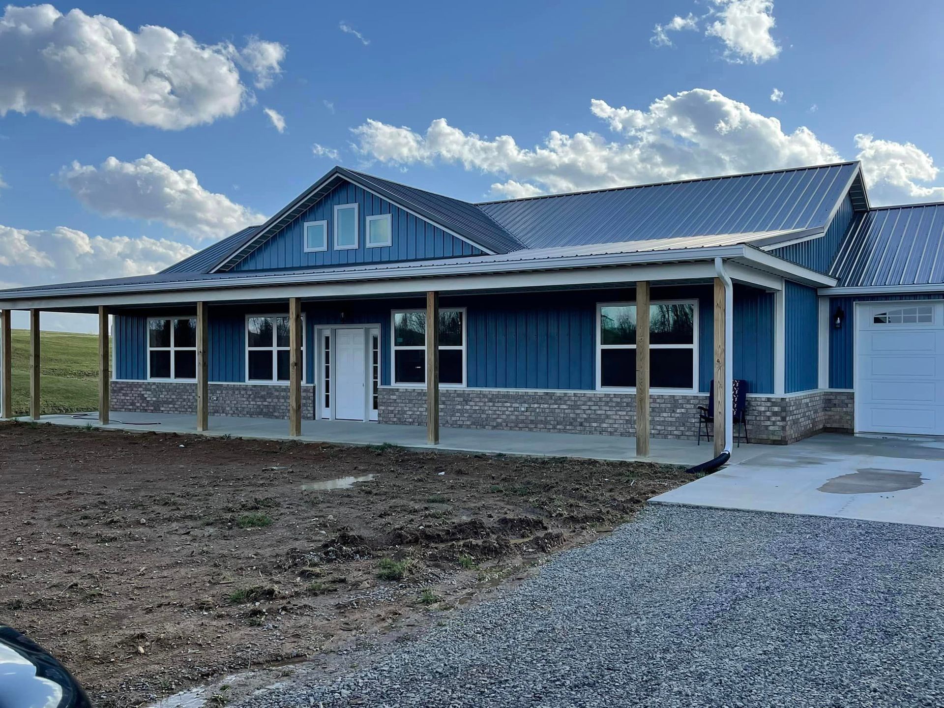 Blue ranch-style house with a large porch and a metal roof under a cloudy sky. Gravel driveway, with front yard under construction.
