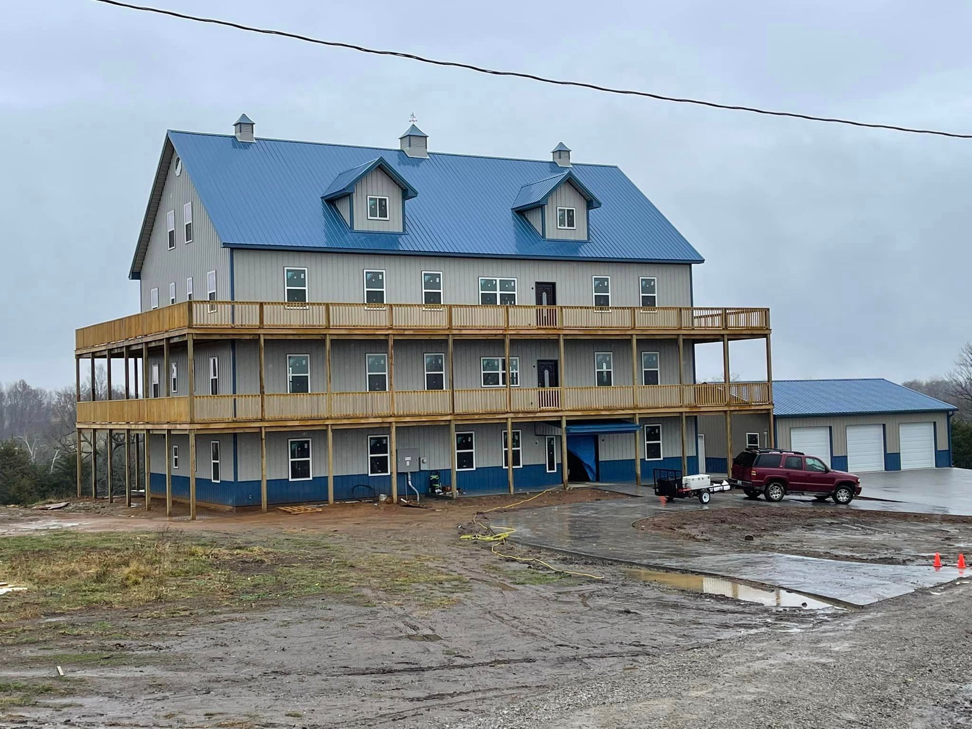 A three-story barn with a blue roof and balconies, under overcast skies, with a pickup truck parked in front.