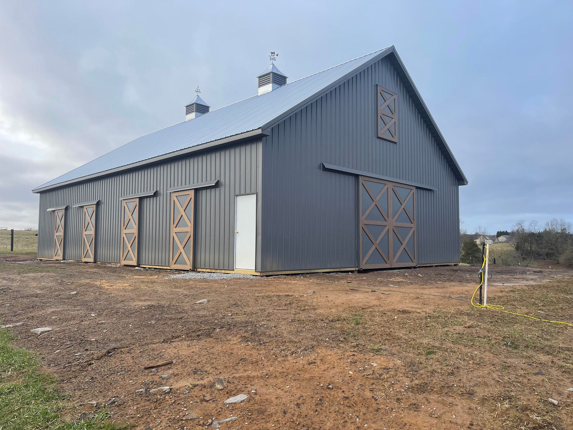 A large, dark grey metal barn with wooden doors and accents stands on a brown, dirt field under a cloudy sky.