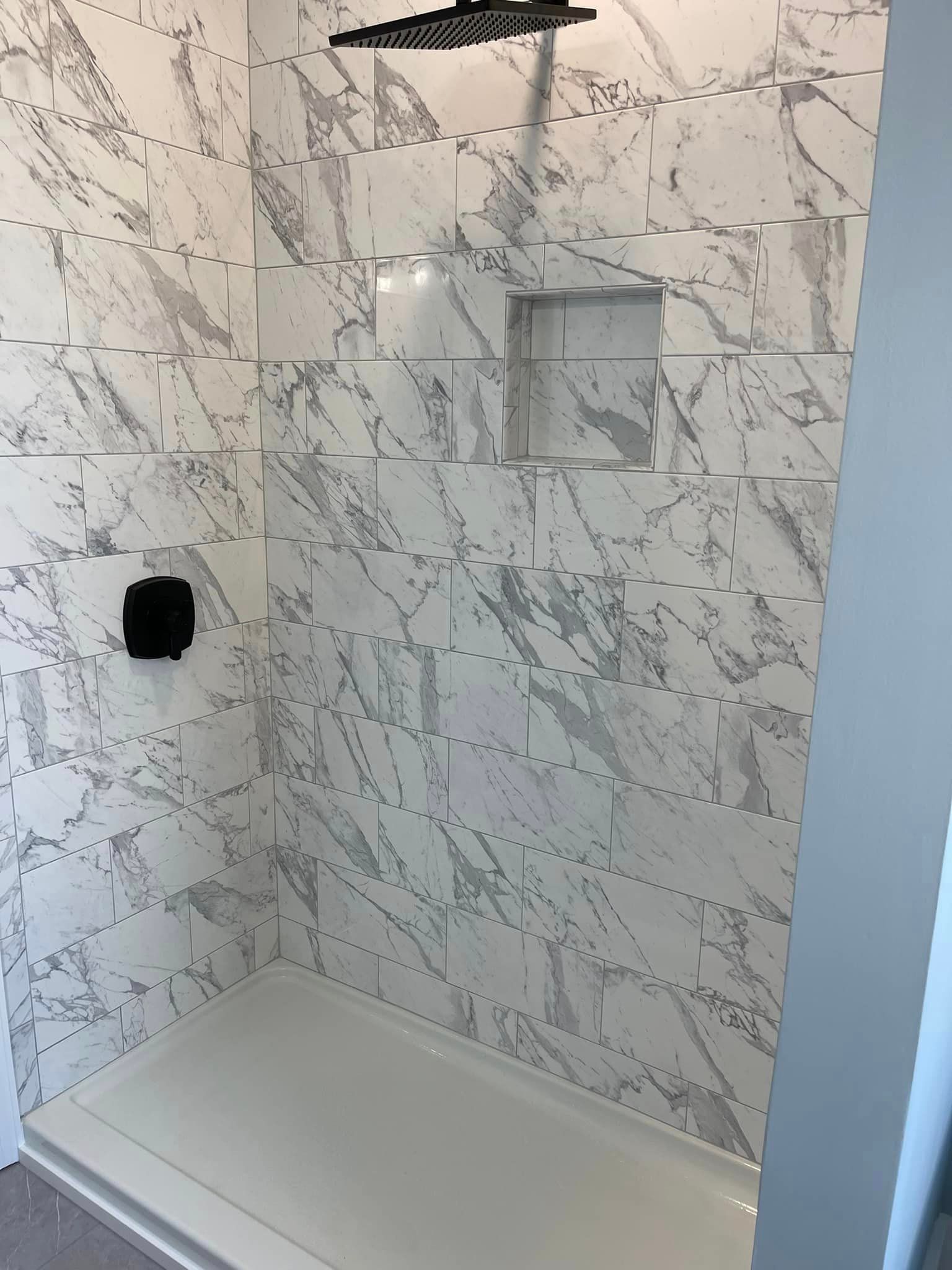 Shower stall with white marble-look tile, black showerhead and controls, and built-in shelf. White shower base.