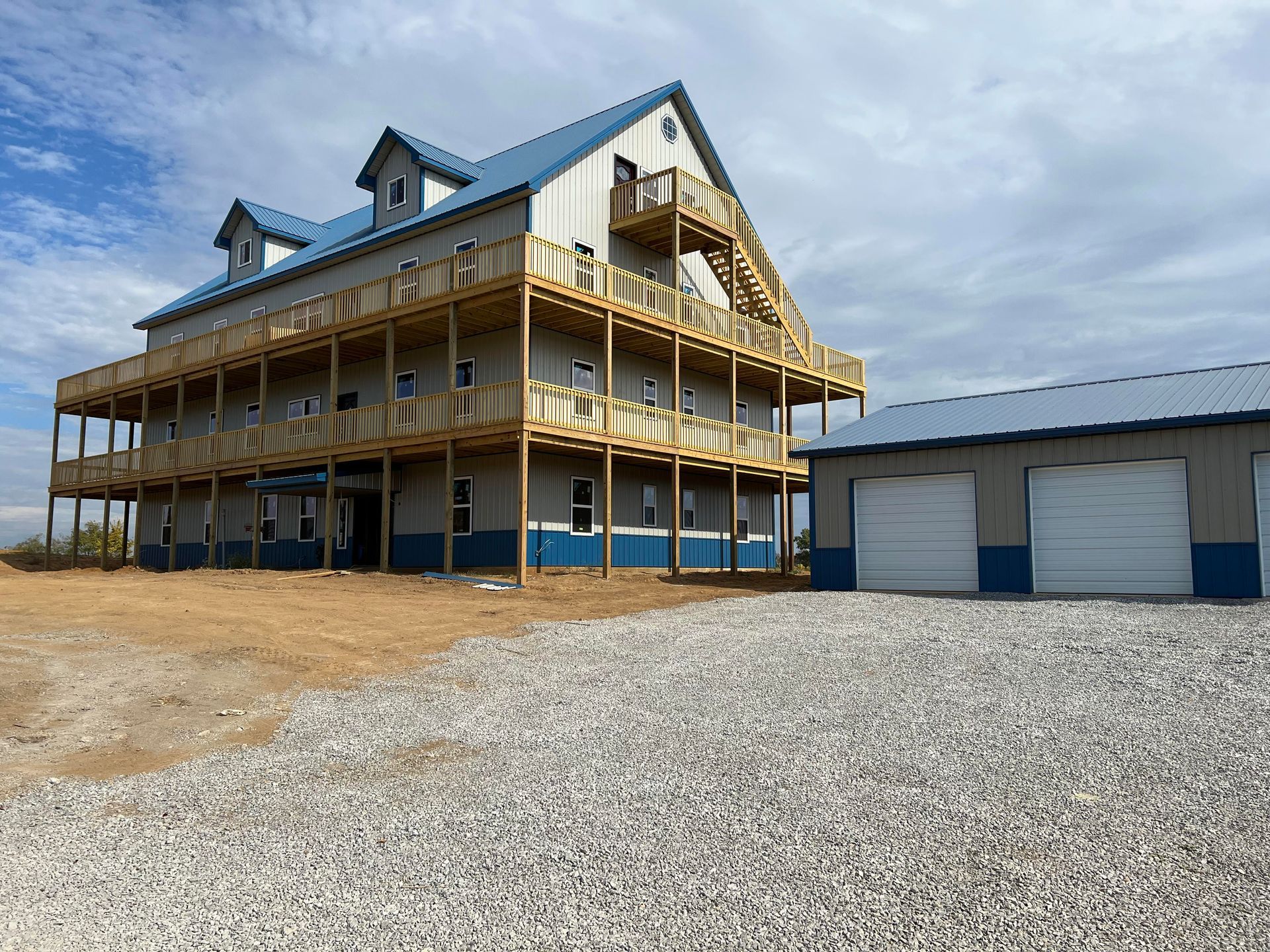 Three-story building with blue roof and decks, next to a smaller garage. Set on gravel with a cloudy sky.