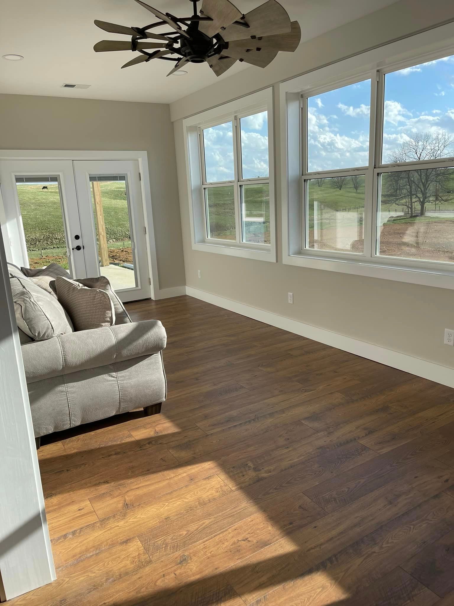 Sunroom with hardwood floors, a gray couch, and large windows overlooking a landscape. A ceiling fan hangs above.