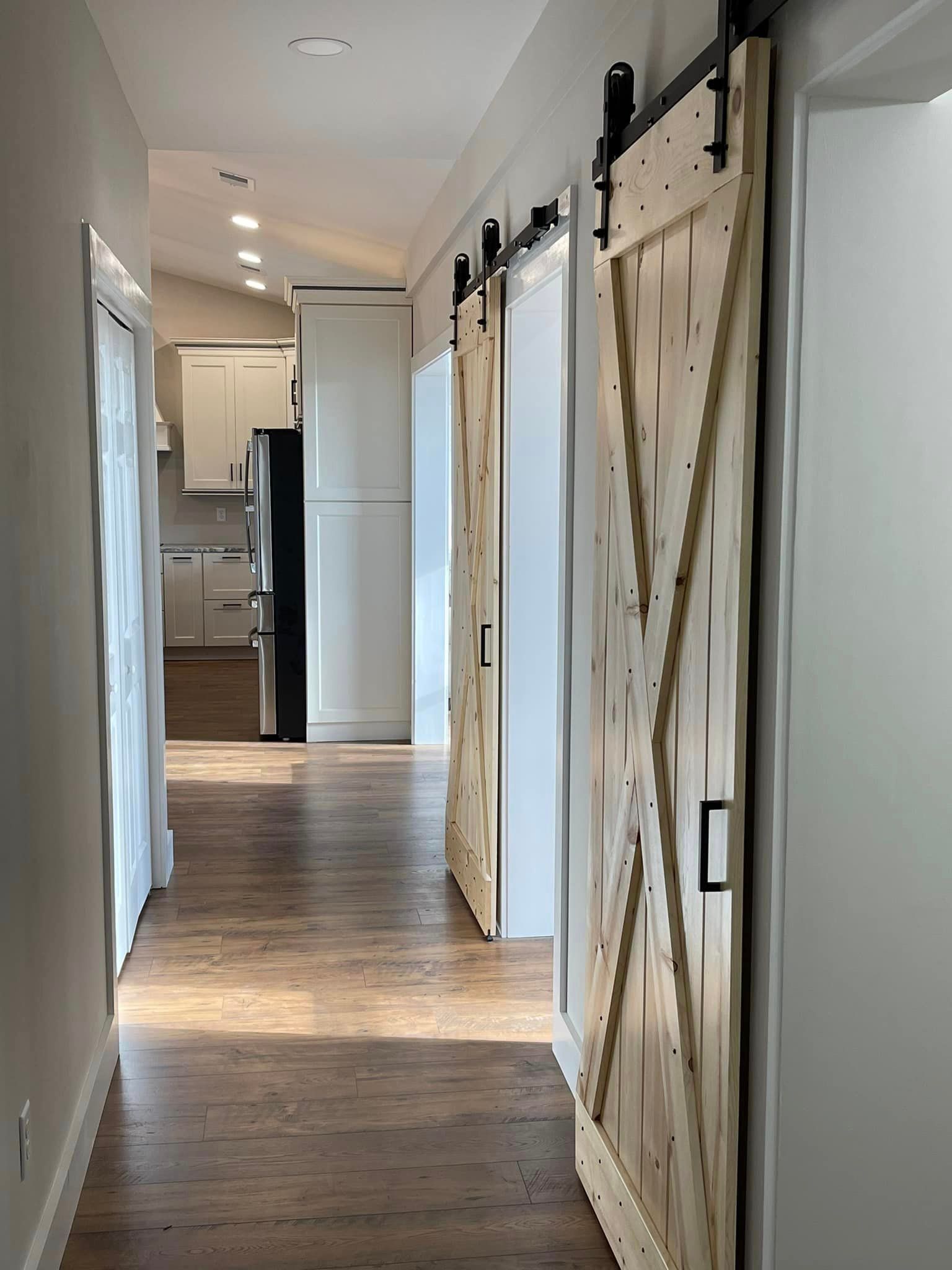 Hallway with wooden sliding barn doors, leading to a kitchen with white cabinets and a black refrigerator.
