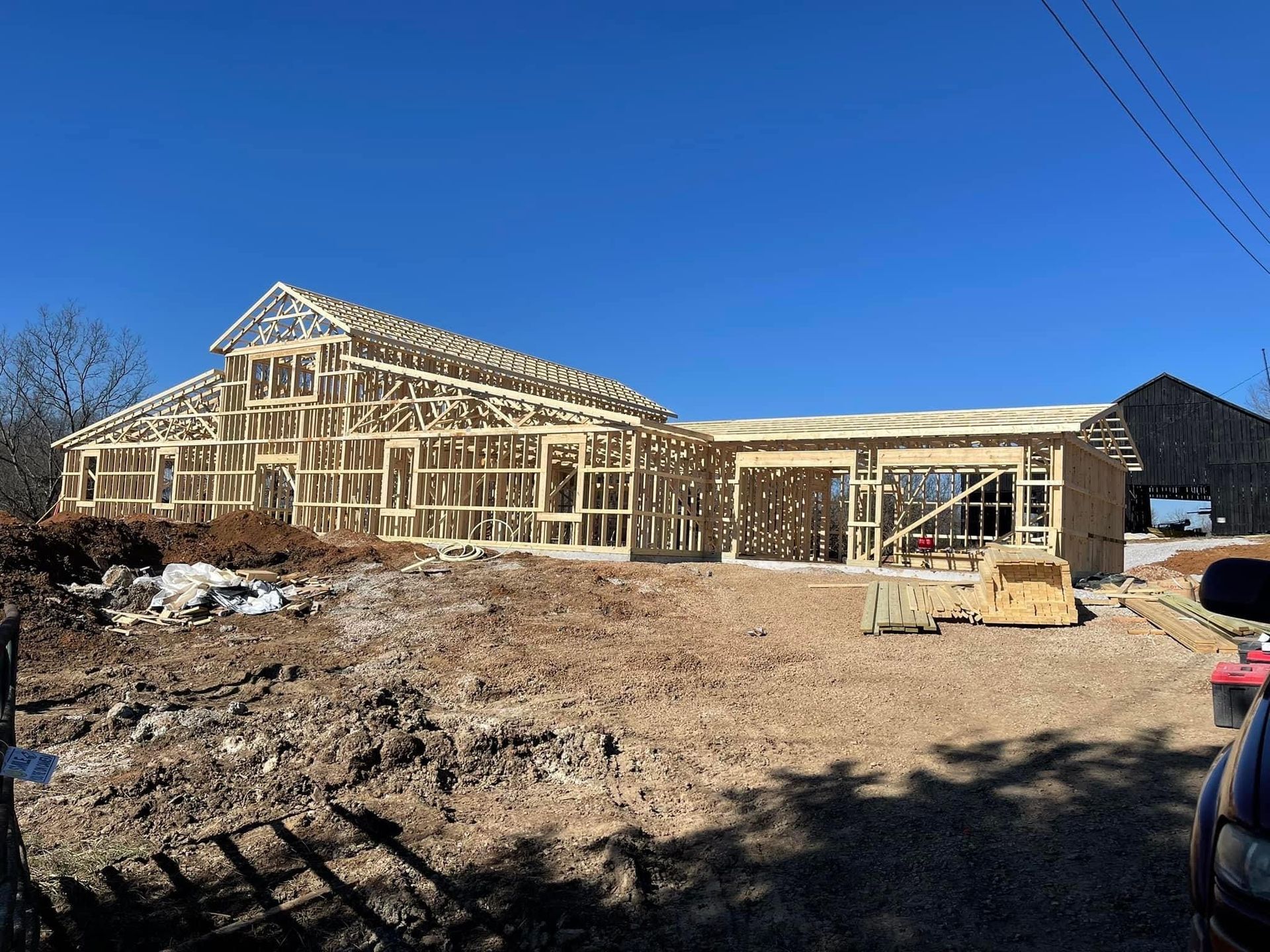 A wooden house under construction on a dirt lot under a clear blue sky. The frame is complete with rafters.