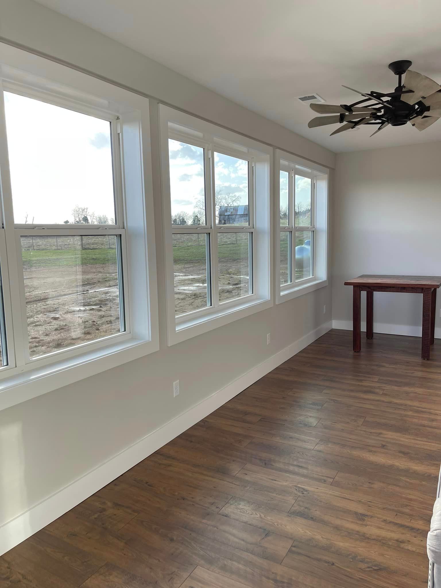 A room with three white-framed windows, a wooden table, and a ceiling fan; hardwood floors and light gray walls.