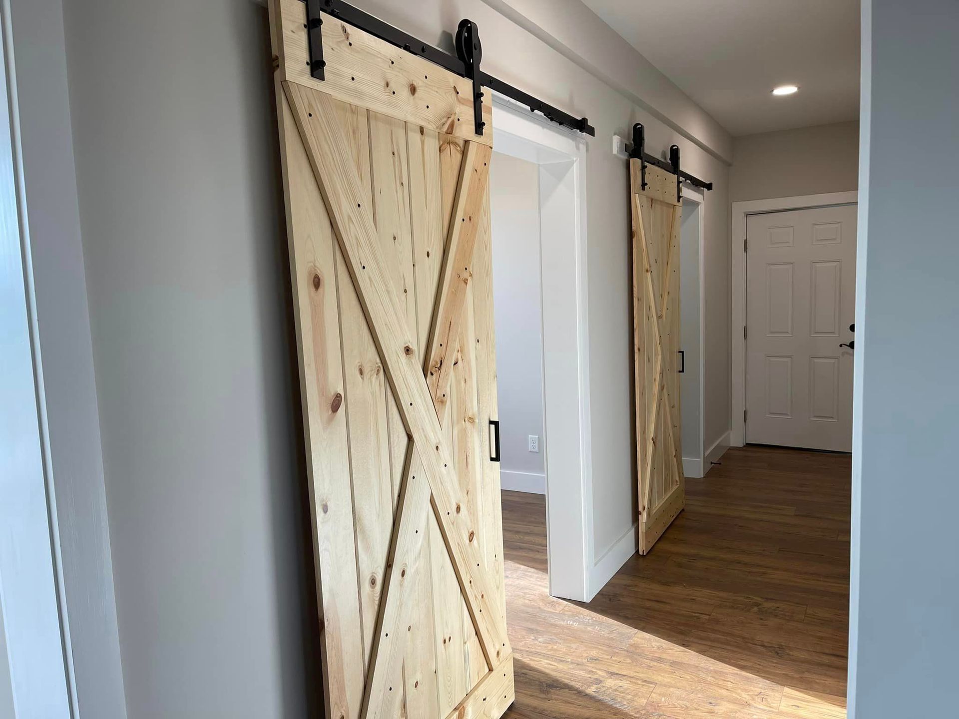 Two light-colored wooden barn doors with black hardware in a hallway. One door is partially open, revealing a room.