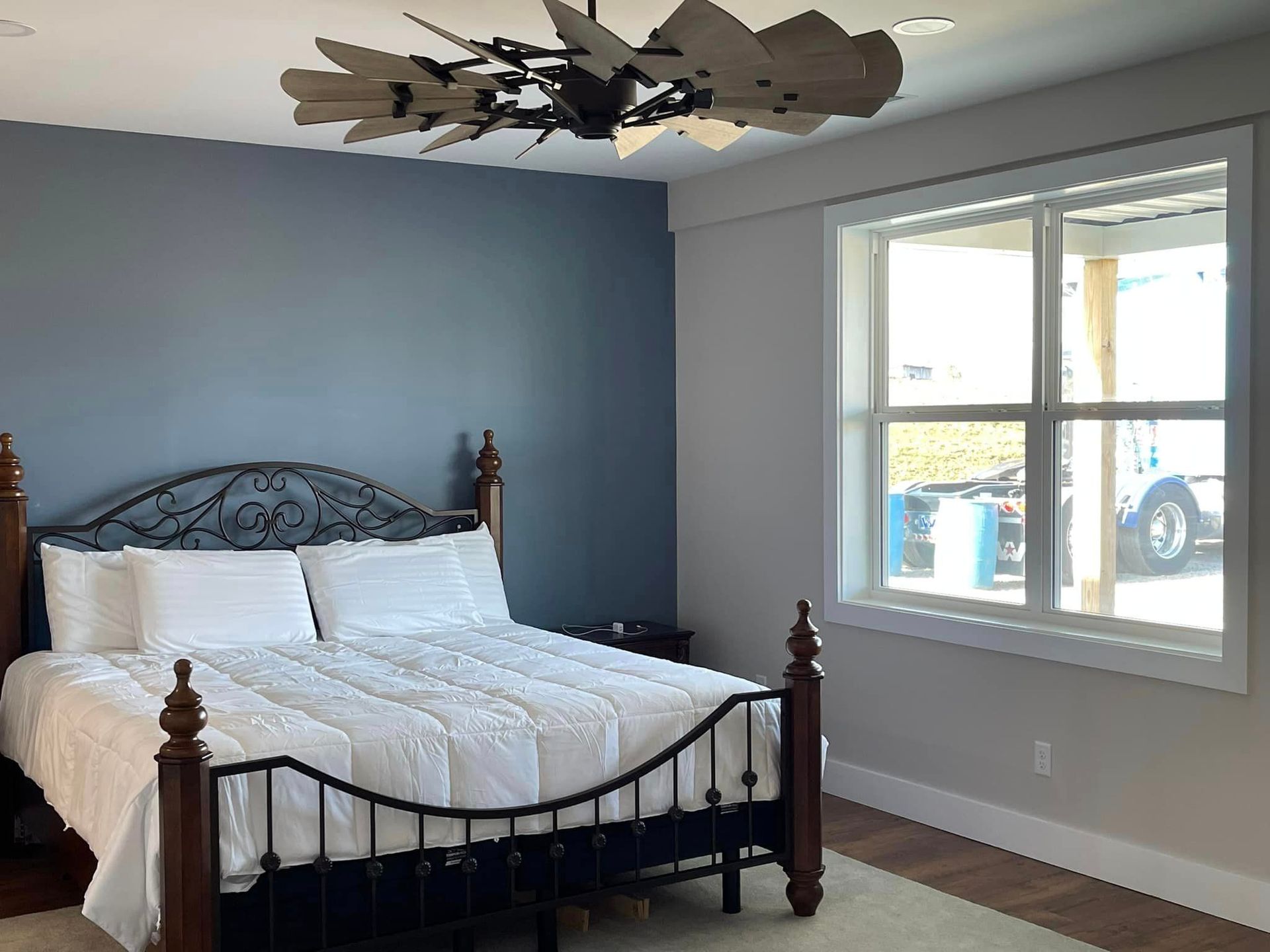 Bedroom with a dark metal bed, white bedding, and a blue wall. A multi-blade fan hangs from the ceiling above the bed.