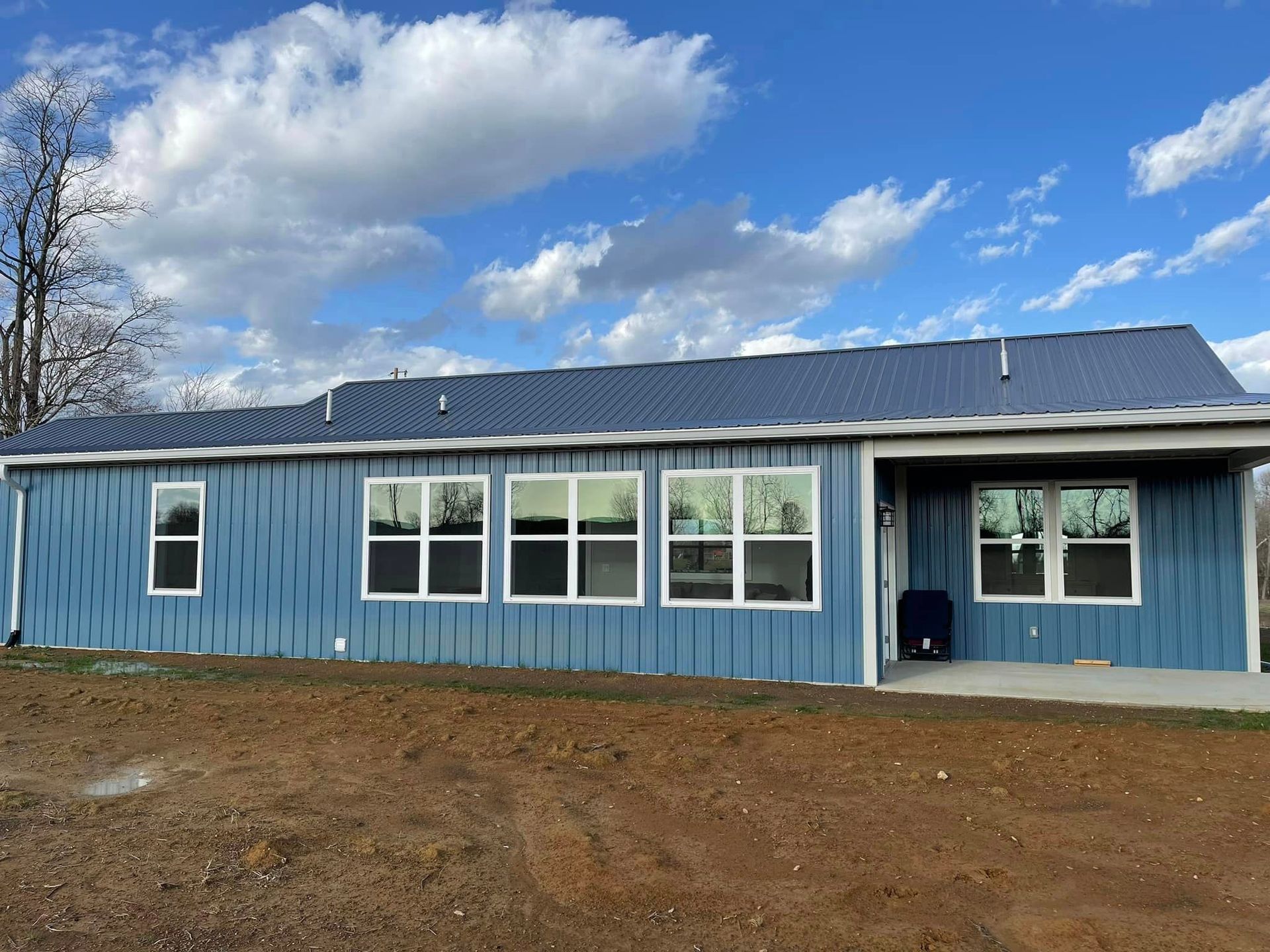 Blue house with metal roof under a partly cloudy sky. The house has multiple windows and sits on a dirt patch.