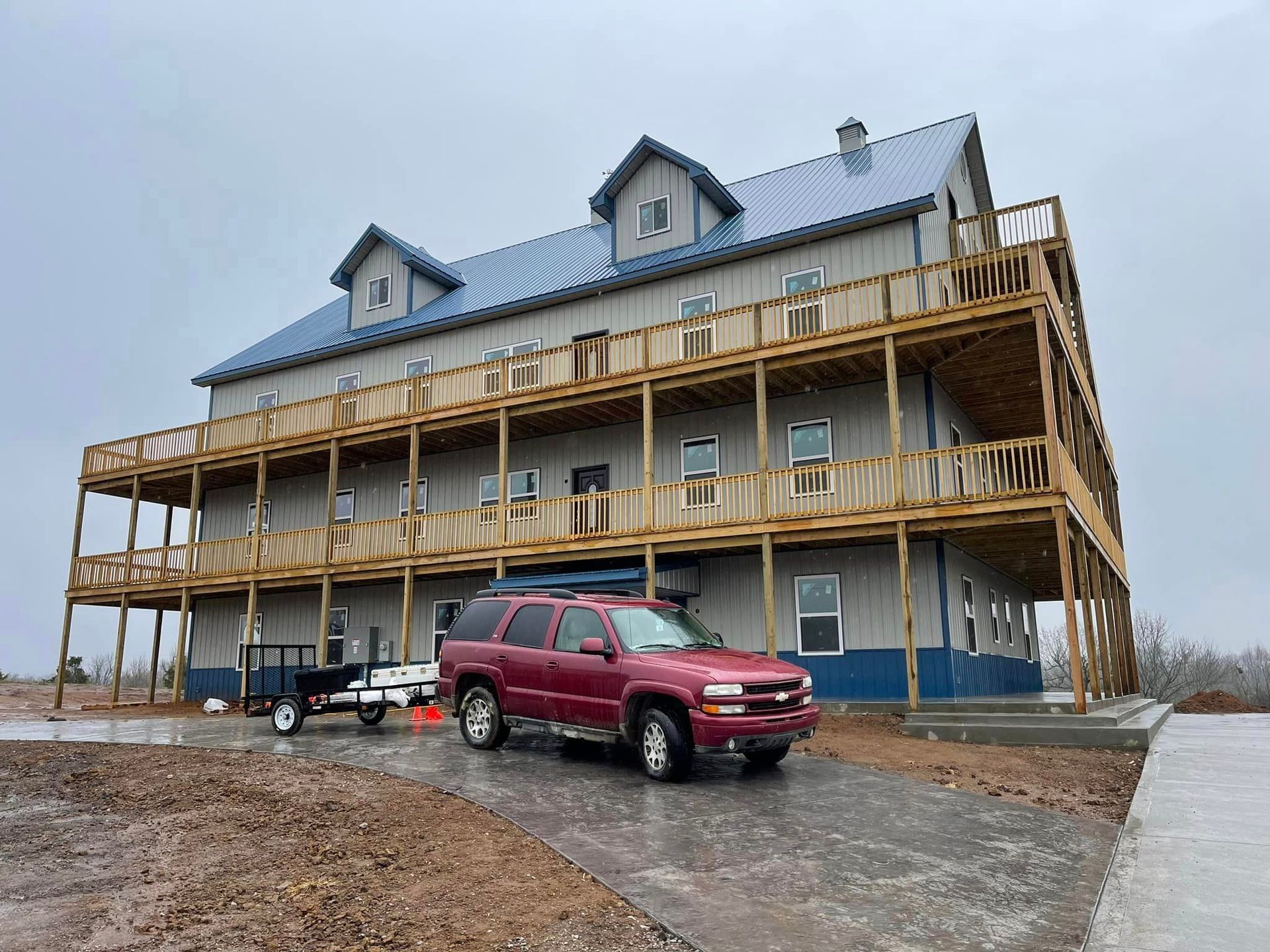 Three-story building with wooden balconies, a blue roof, and a red SUV parked in front on a wet, overcast day.