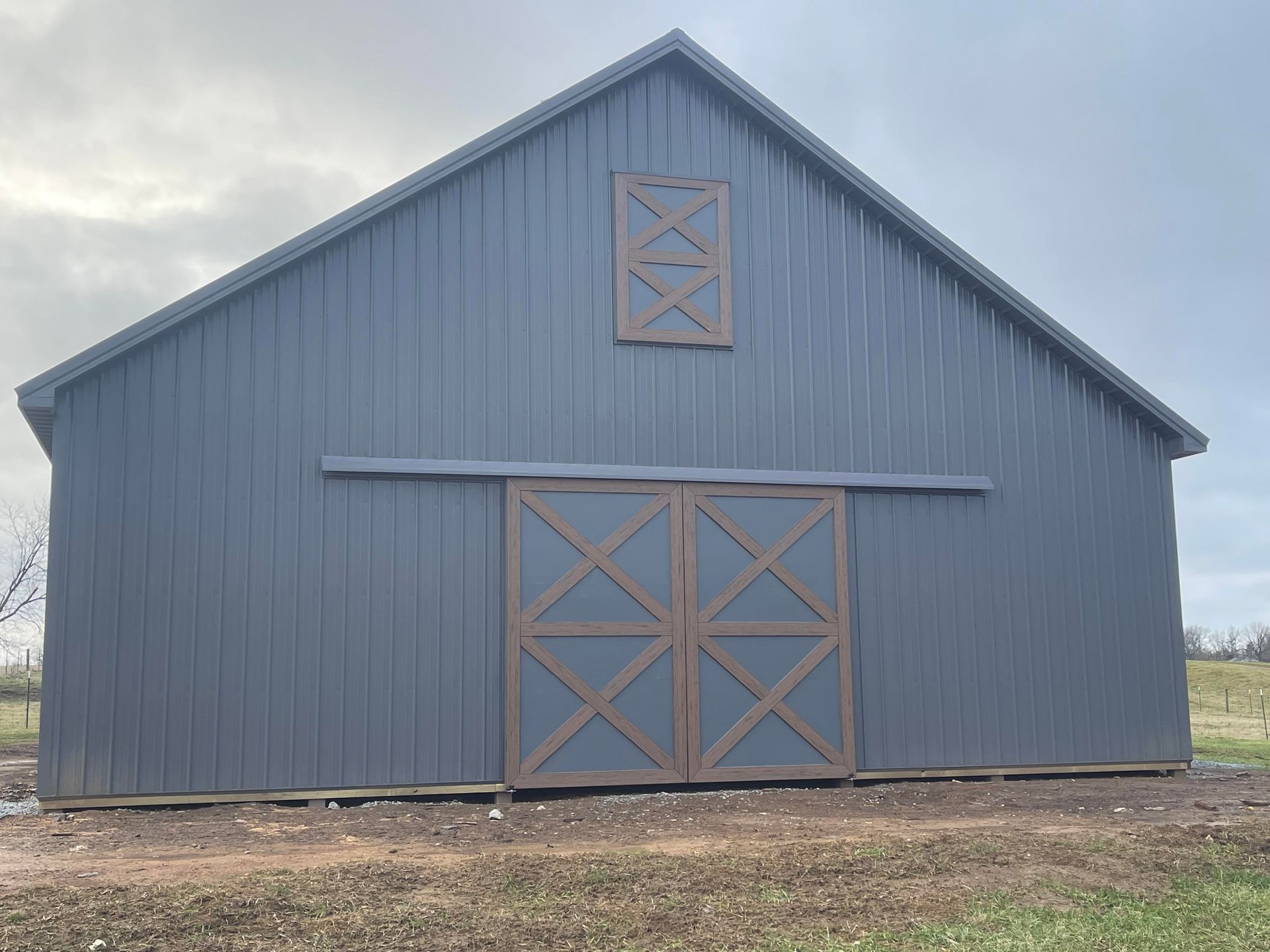 Gray metal barn with brown wooden doors and a window, set against a cloudy sky.