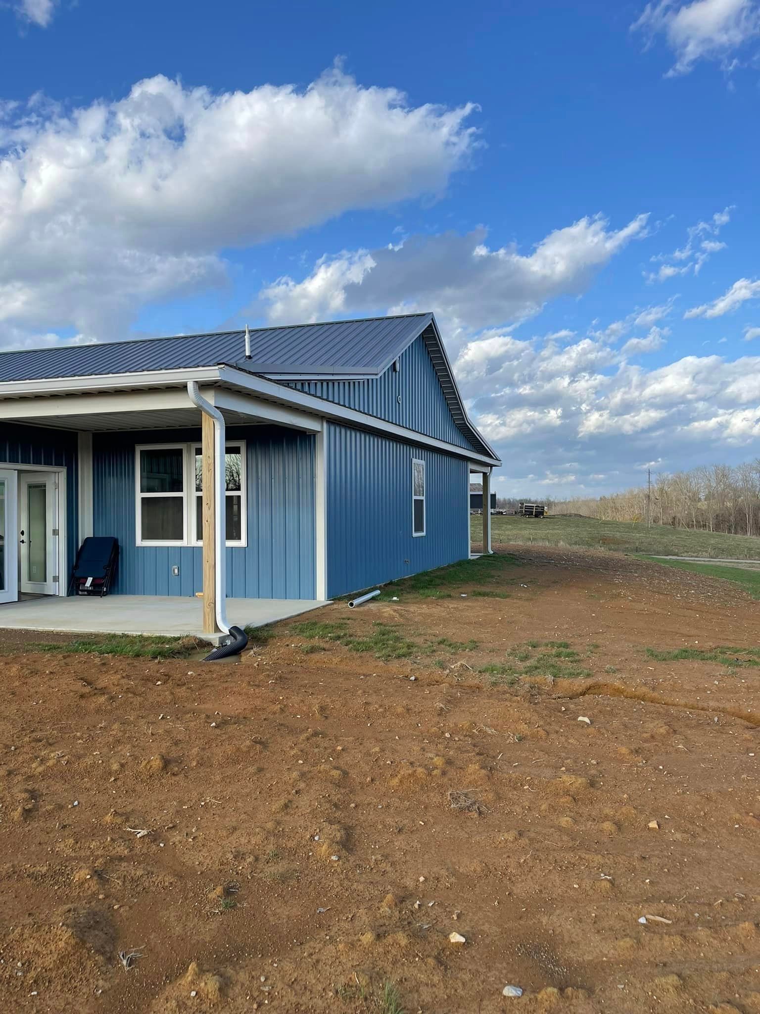 Blue house with porch and dark blue roof against a blue sky with scattered clouds. The building sits on a brown, unpaved lot.