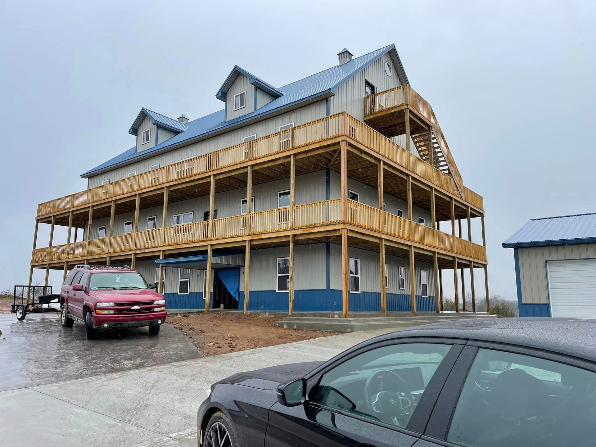 Three-story gray building with blue trim and a wrap-around wooden deck; a red SUV and black car are parked in front.