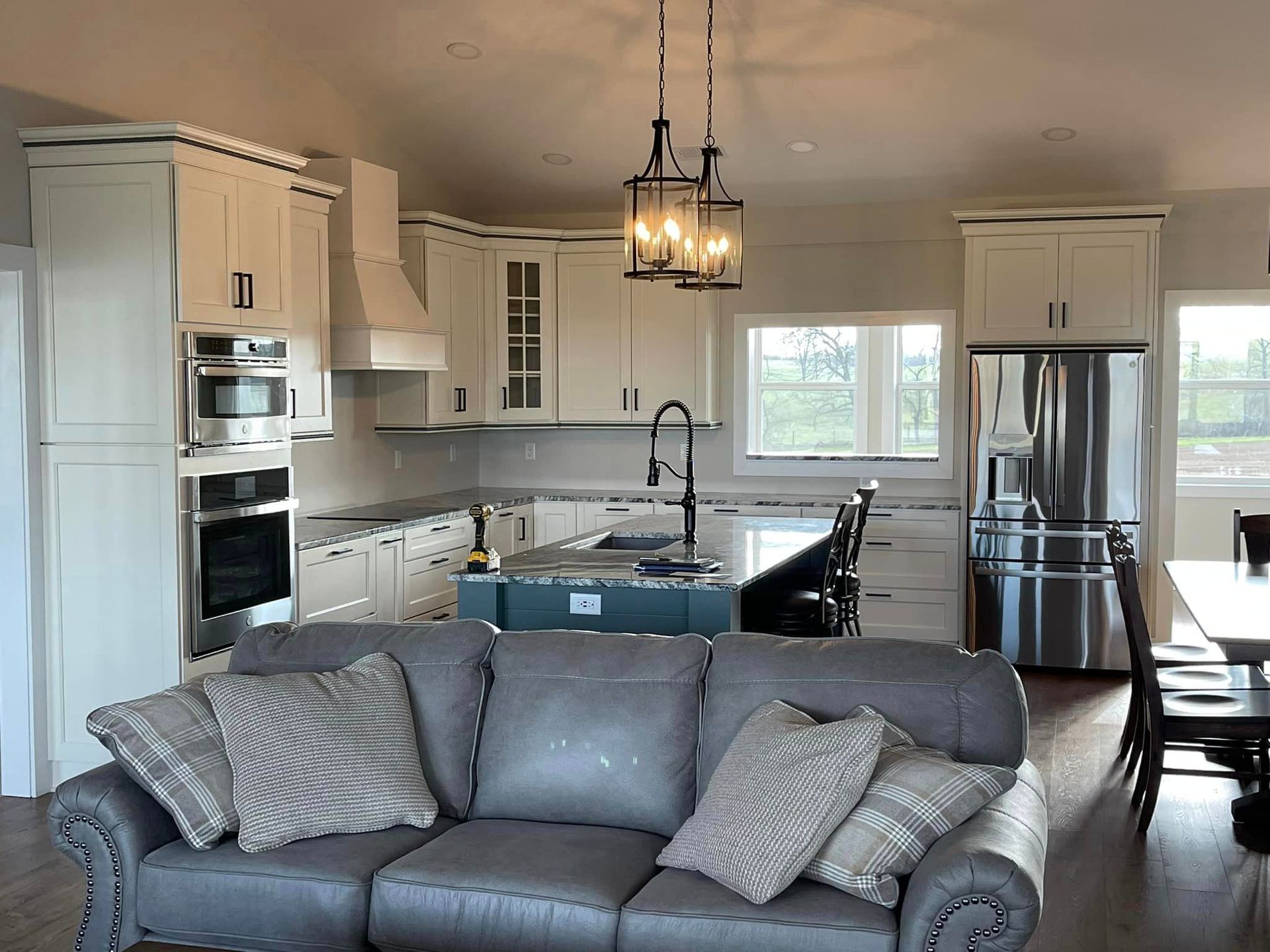 A spacious kitchen with white cabinets, stainless steel appliances, and a blue island is viewed from a gray sofa. A lantern-style light hangs above.