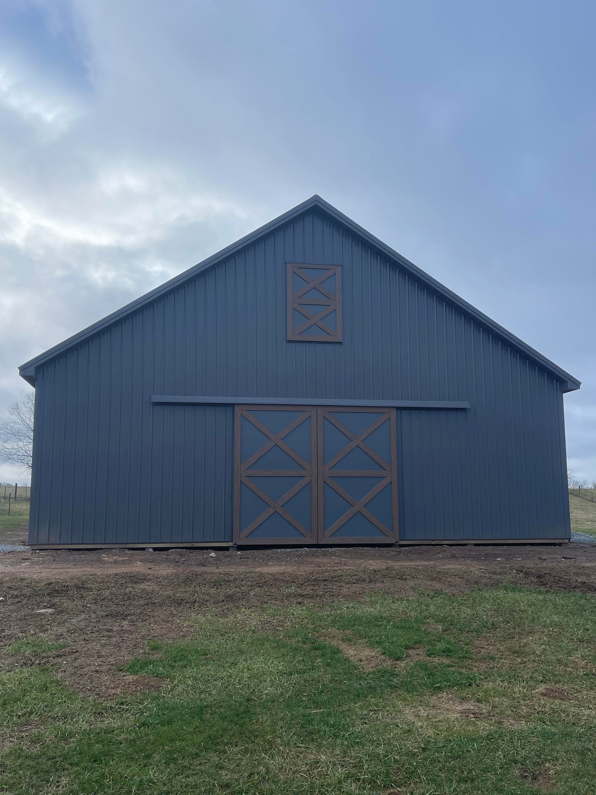 Gray metal barn with brown sliding doors and a smaller decorative window. The barn sits on a grassy field under a cloudy sky.