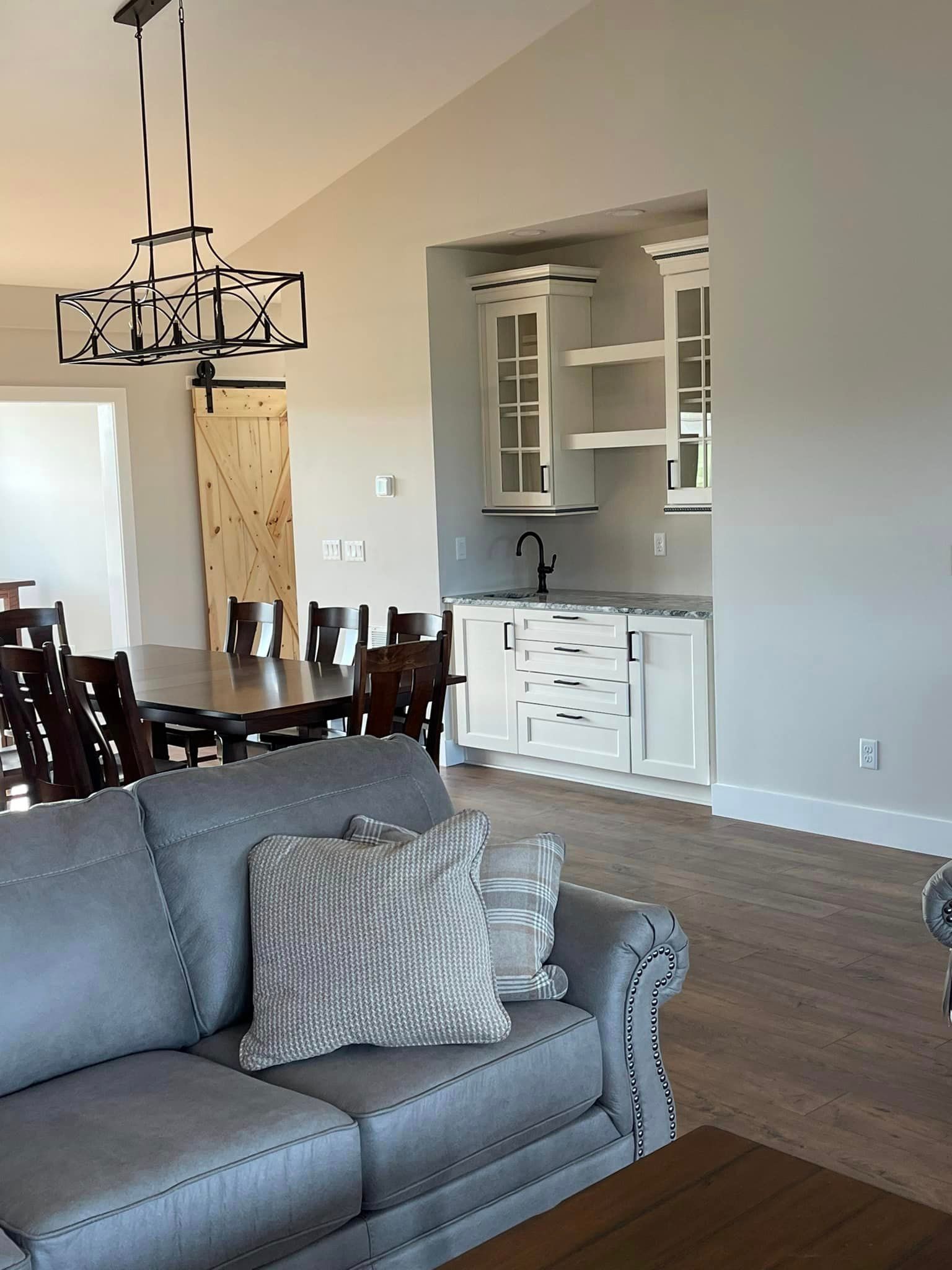 A modern living space with a gray sofa, dining table, and built-in bar area with white cabinets. A barn door is visible.