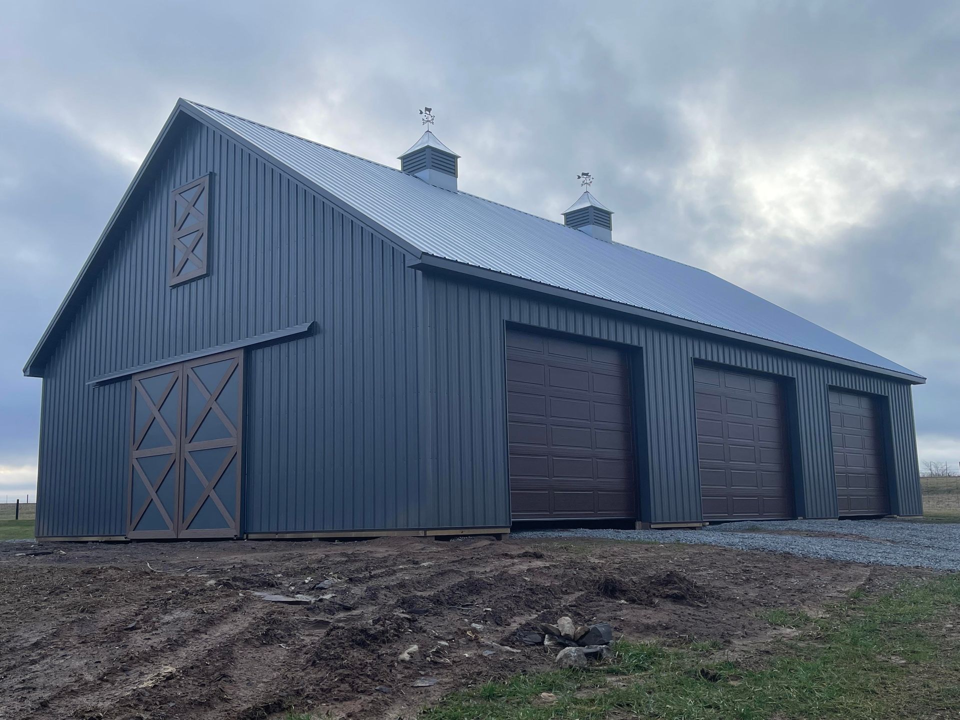 A large gray barn with a metal roof and three brown garage doors under a cloudy sky.