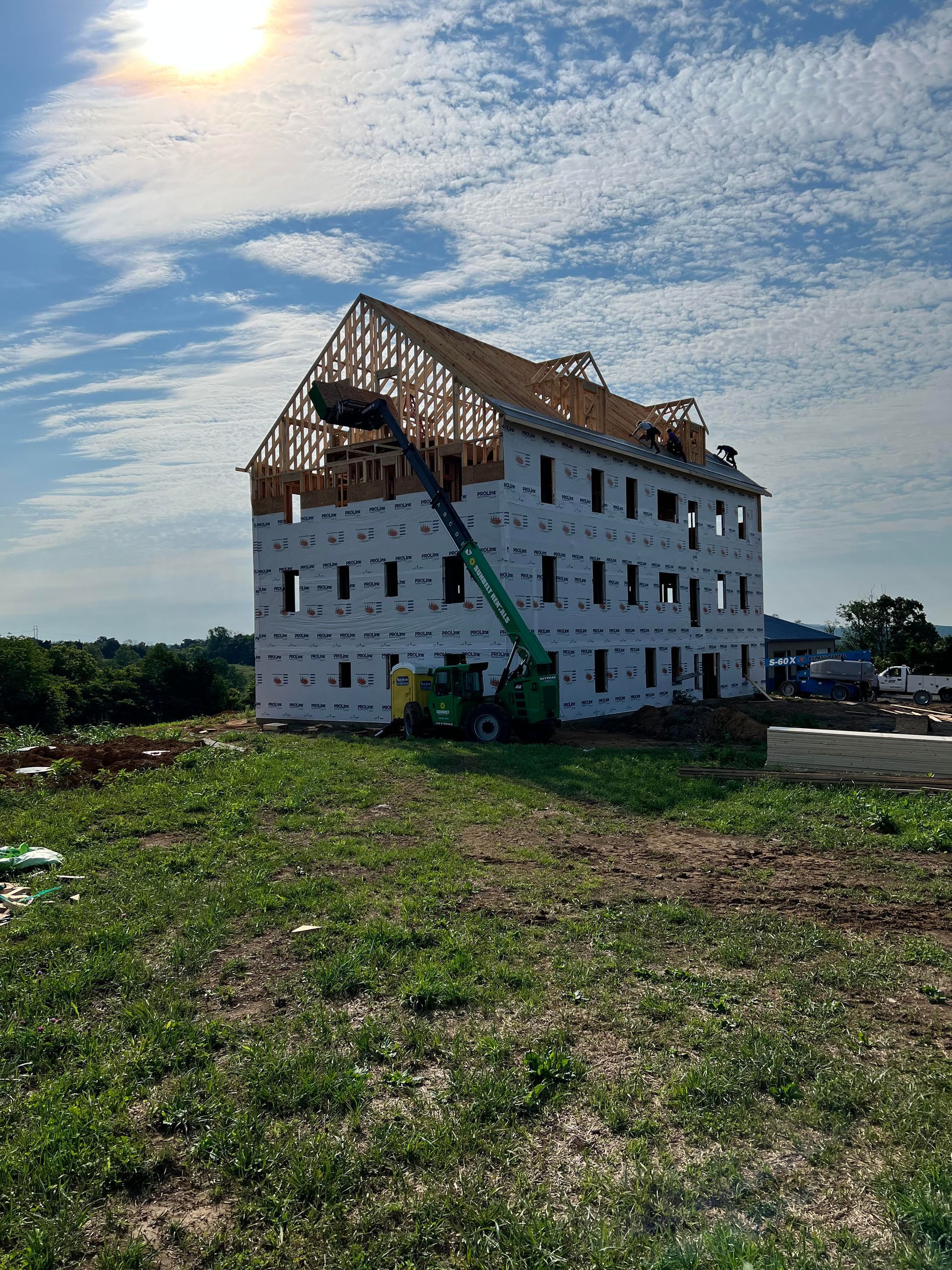A multi-story building under construction with wooden roof framing, a green lift, and a sunny sky. It’s in a grassy field.
