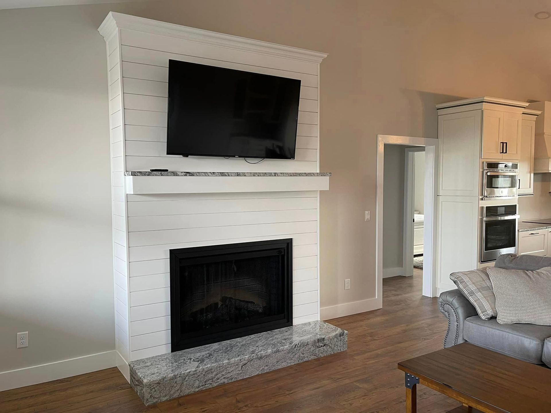 White-paneled fireplace with a black television mounted above the hearth in a living room. Granite base, wood floors.