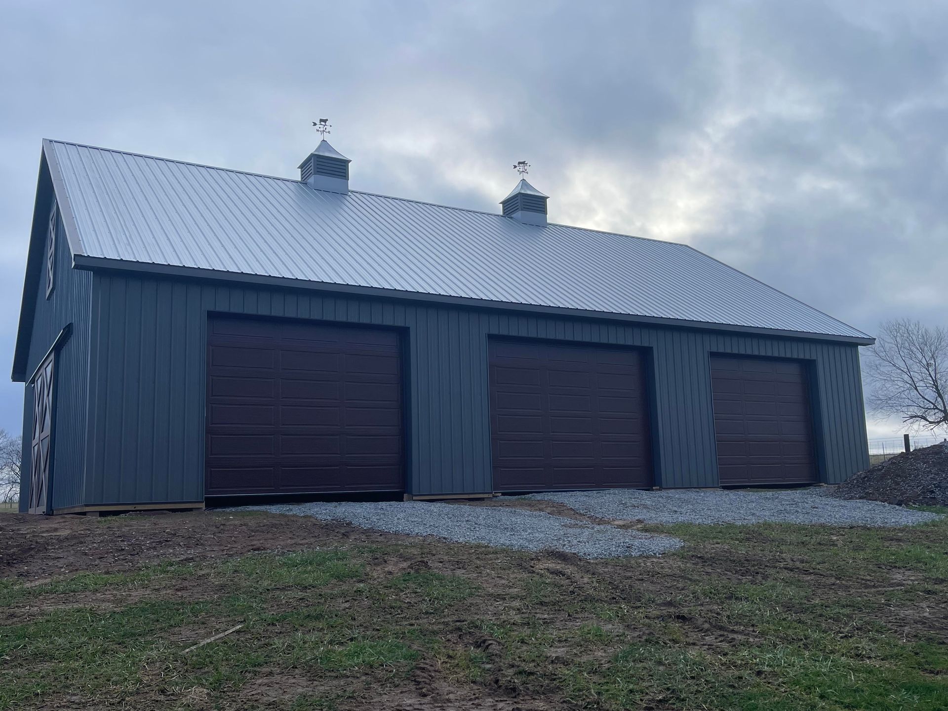 Three-bay garage with gray-blue siding, brown garage doors, and a metal roof. Two cupolas are atop the roof under an overcast sky.