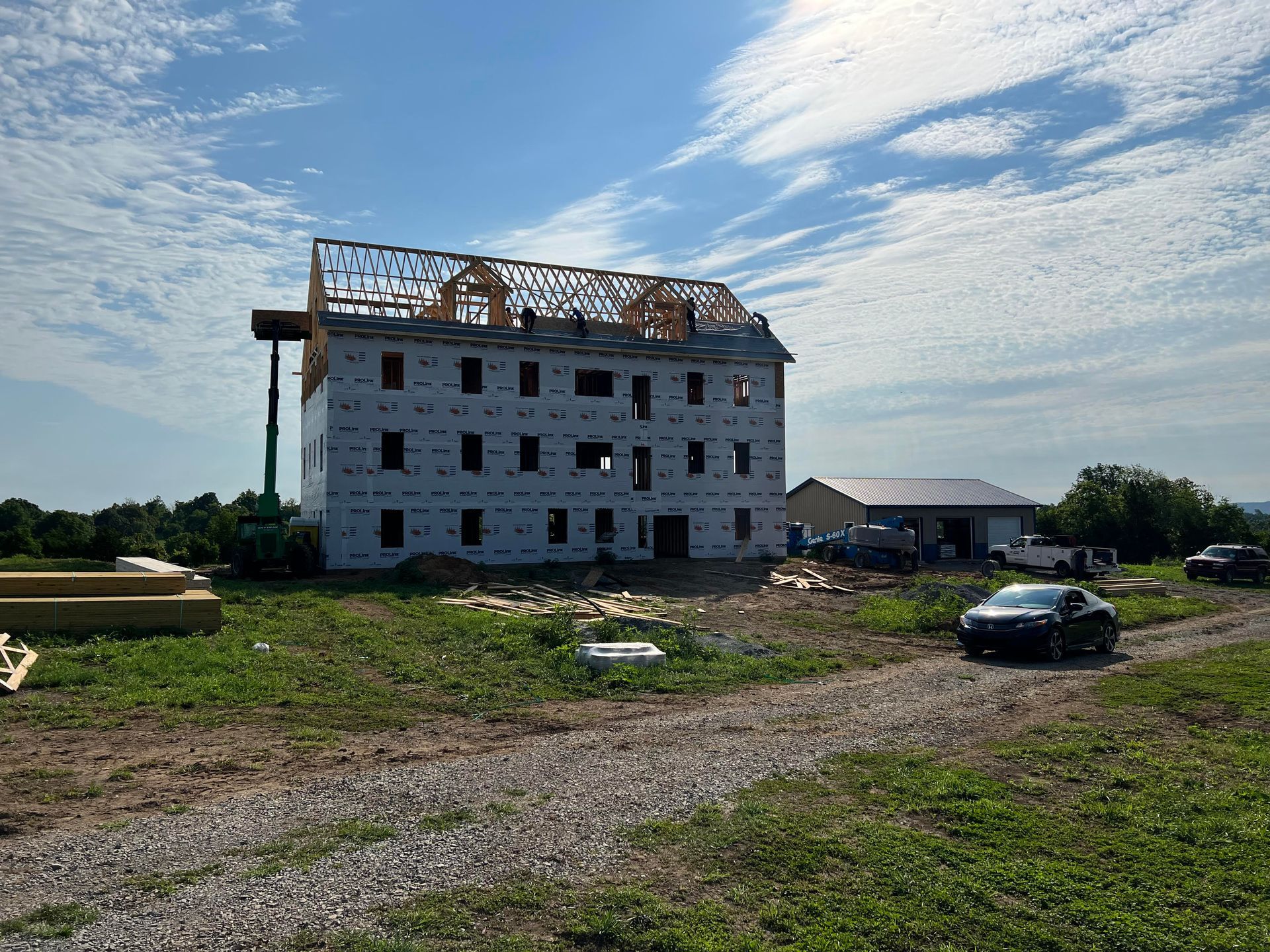 A multi-story building under construction on a grassy lot under a blue sky. The roof framing is visible, and a black car is parked nearby.