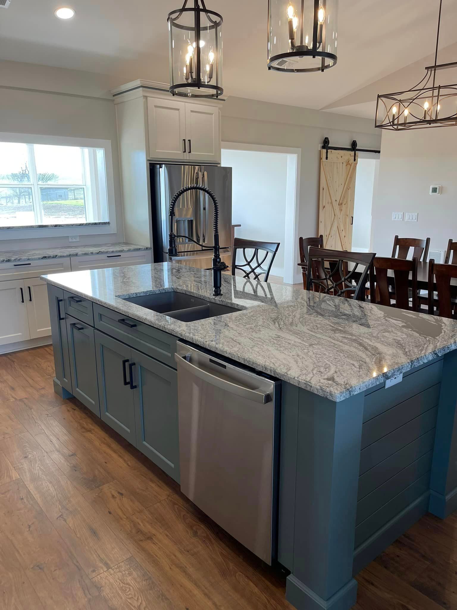 A modern kitchen with a blue island, stainless steel appliances, and granite countertops. Light fixtures hang above the island and dining area.