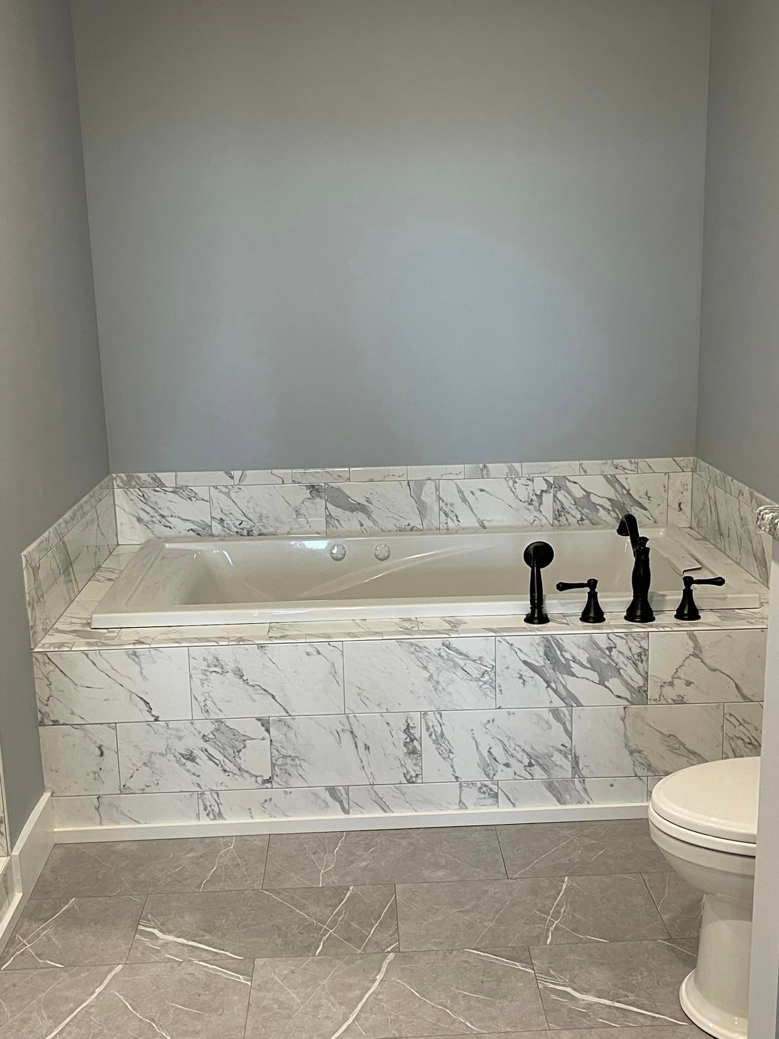 Bathroom with a white marble-look tiled tub and surrounding area. Black fixtures and a toilet are visible. Gray walls and flooring.