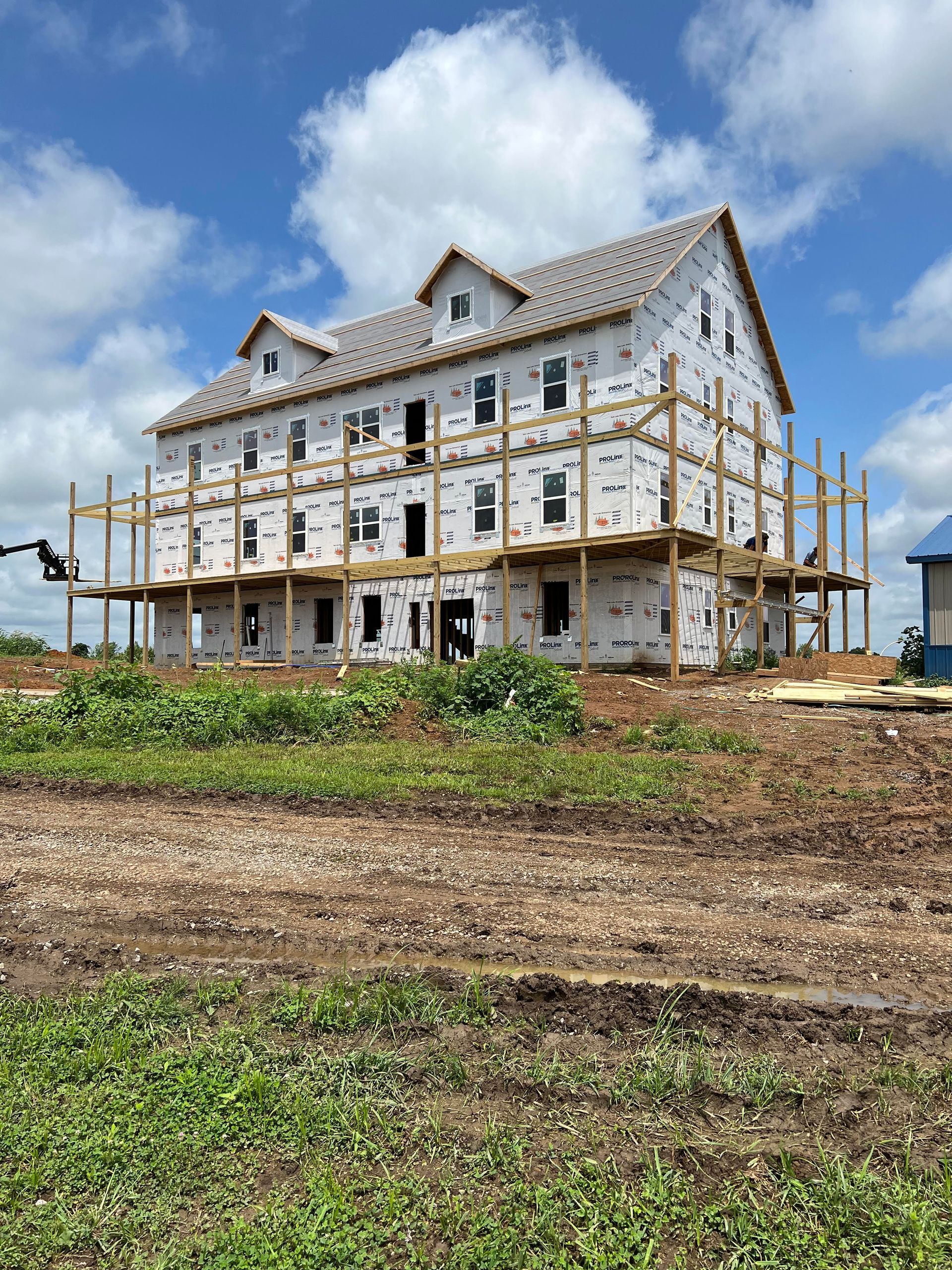 A three-story building under construction wrapped in white material, with wooden scaffolding against a bright blue sky.