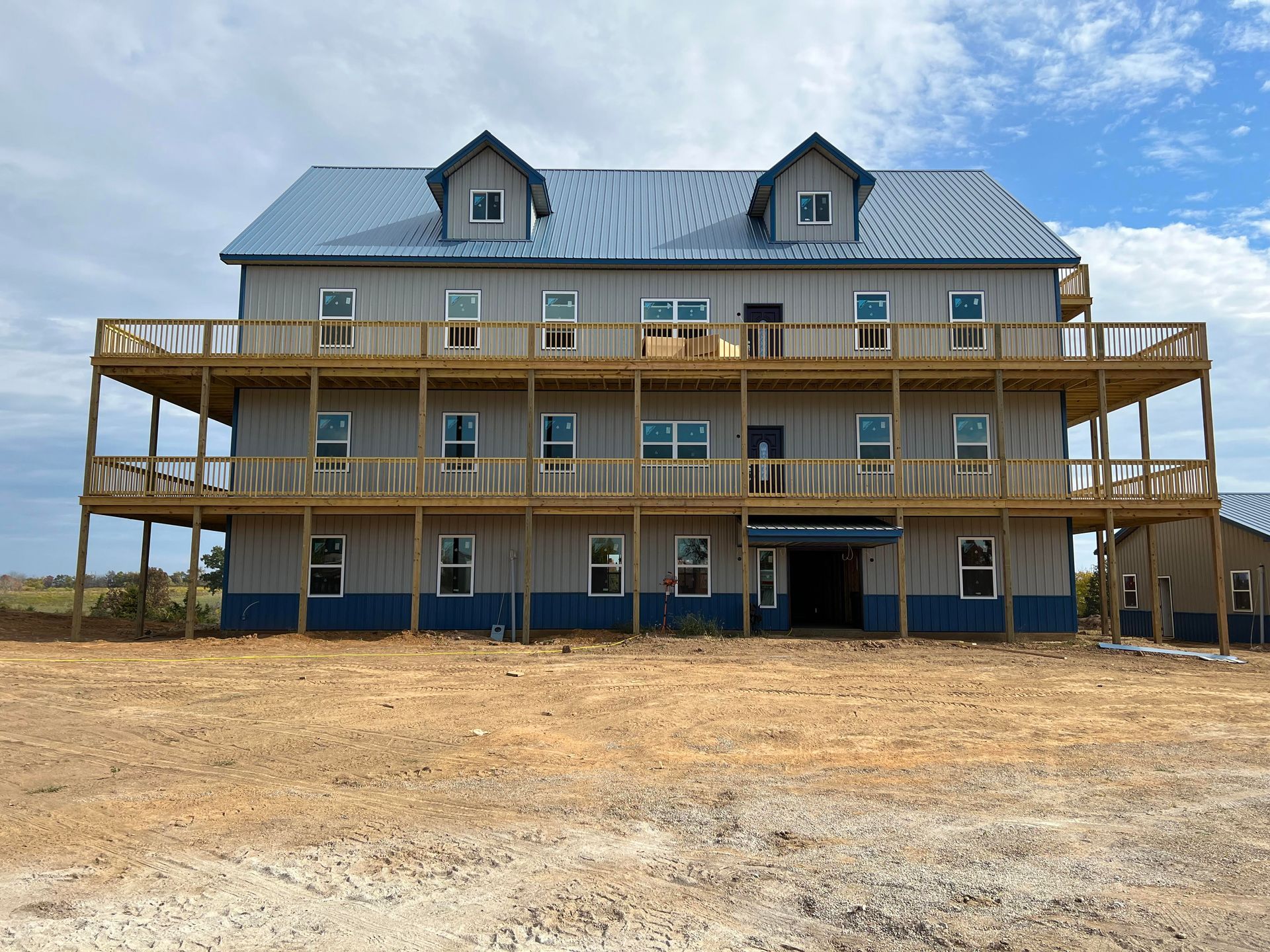 Three-story metal-sided building with wooden balconies and blue roof against a cloudy sky. The building sits on a dirt lot.