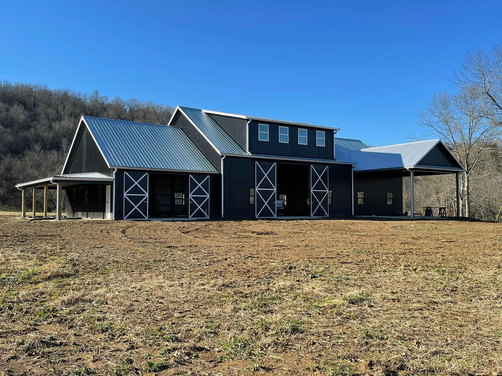 Black barn with white trim and a silver roof under a clear blue sky, in a field with a treeline in the background.