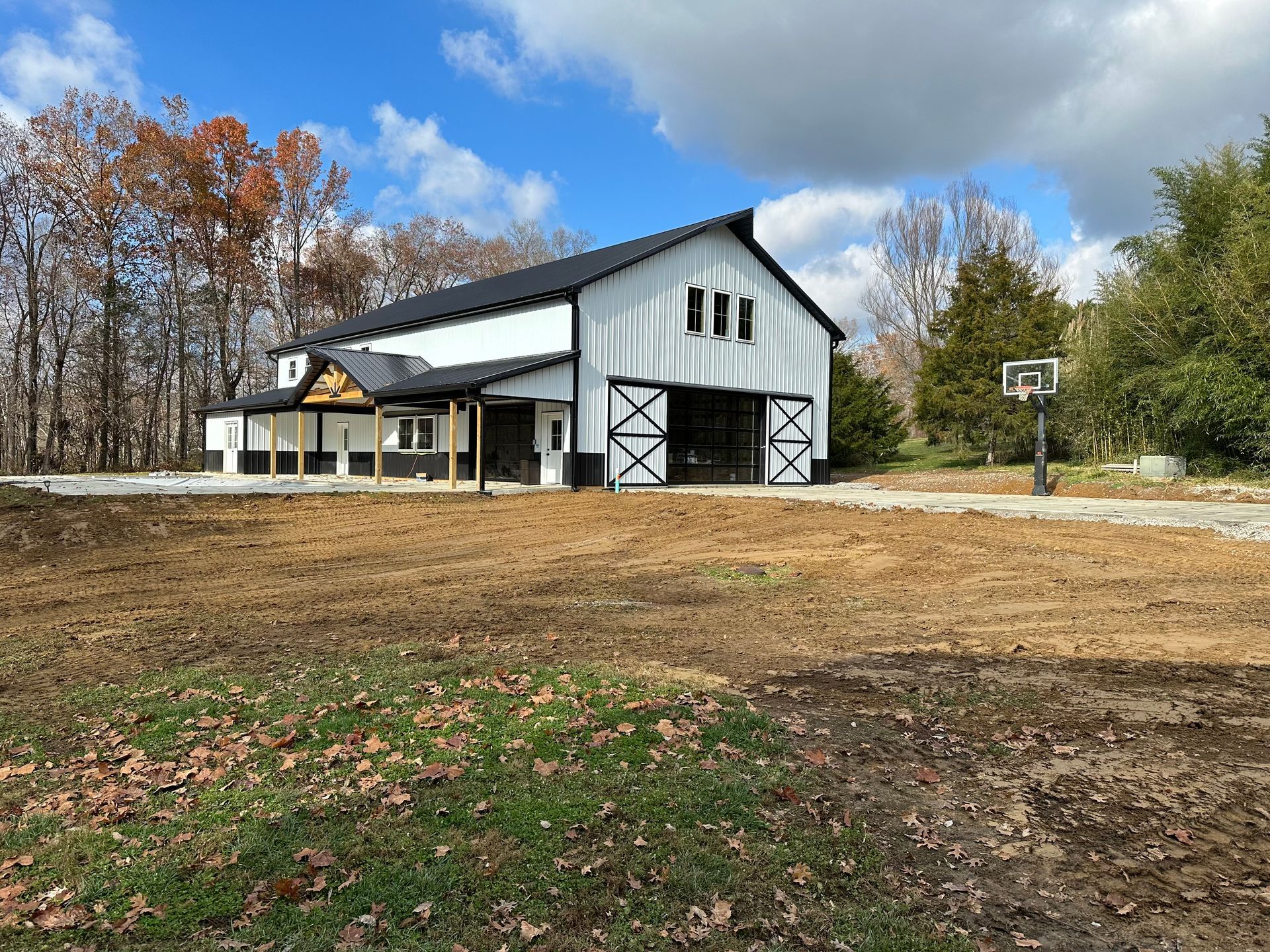 White barn with black trim and large doors, with a porch and basketball hoop in a rural setting under a cloudy sky.