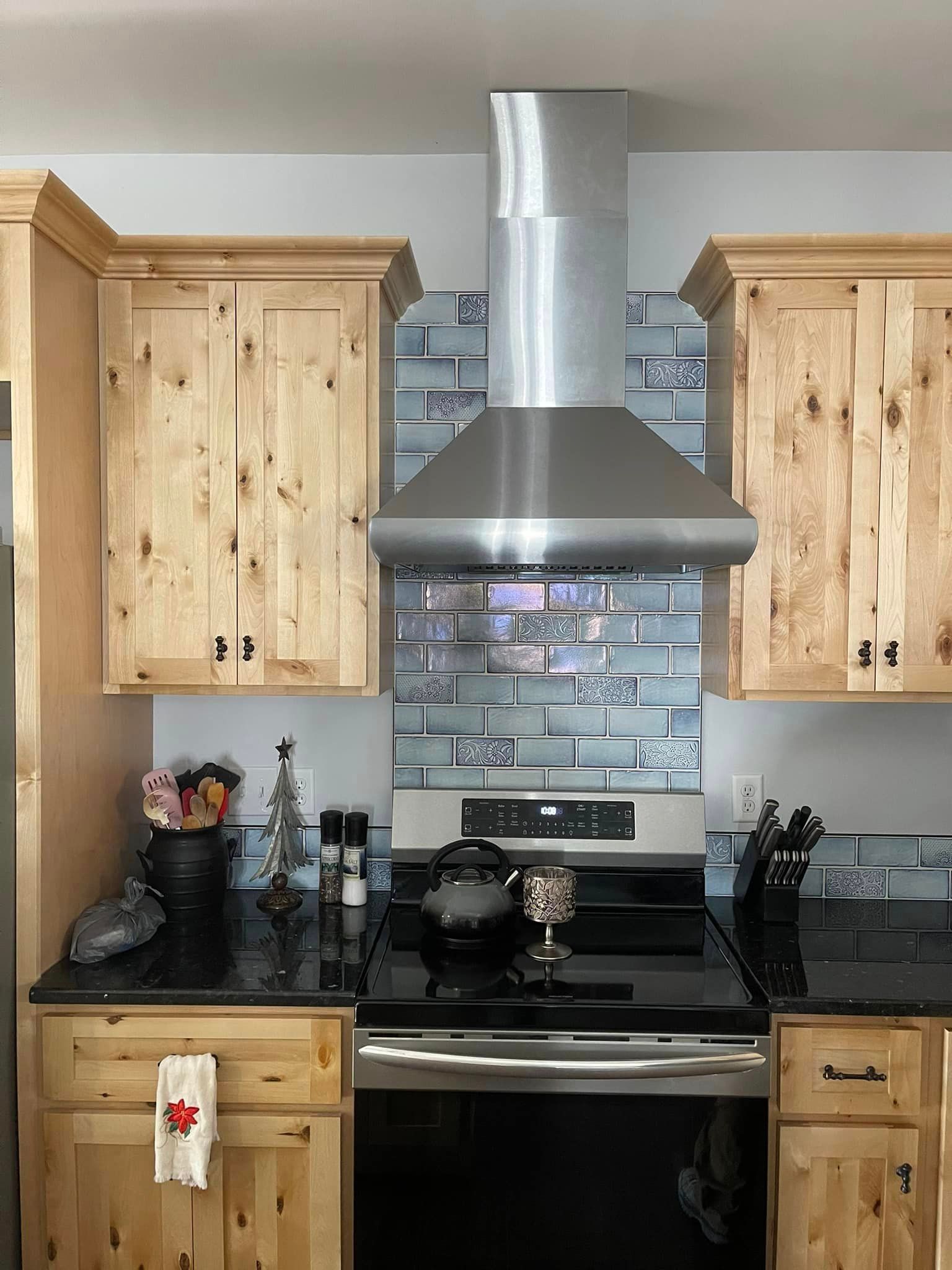 Kitchen with light wood cabinets, stainless steel range hood over a stove with a blue-gray tiled backsplash.