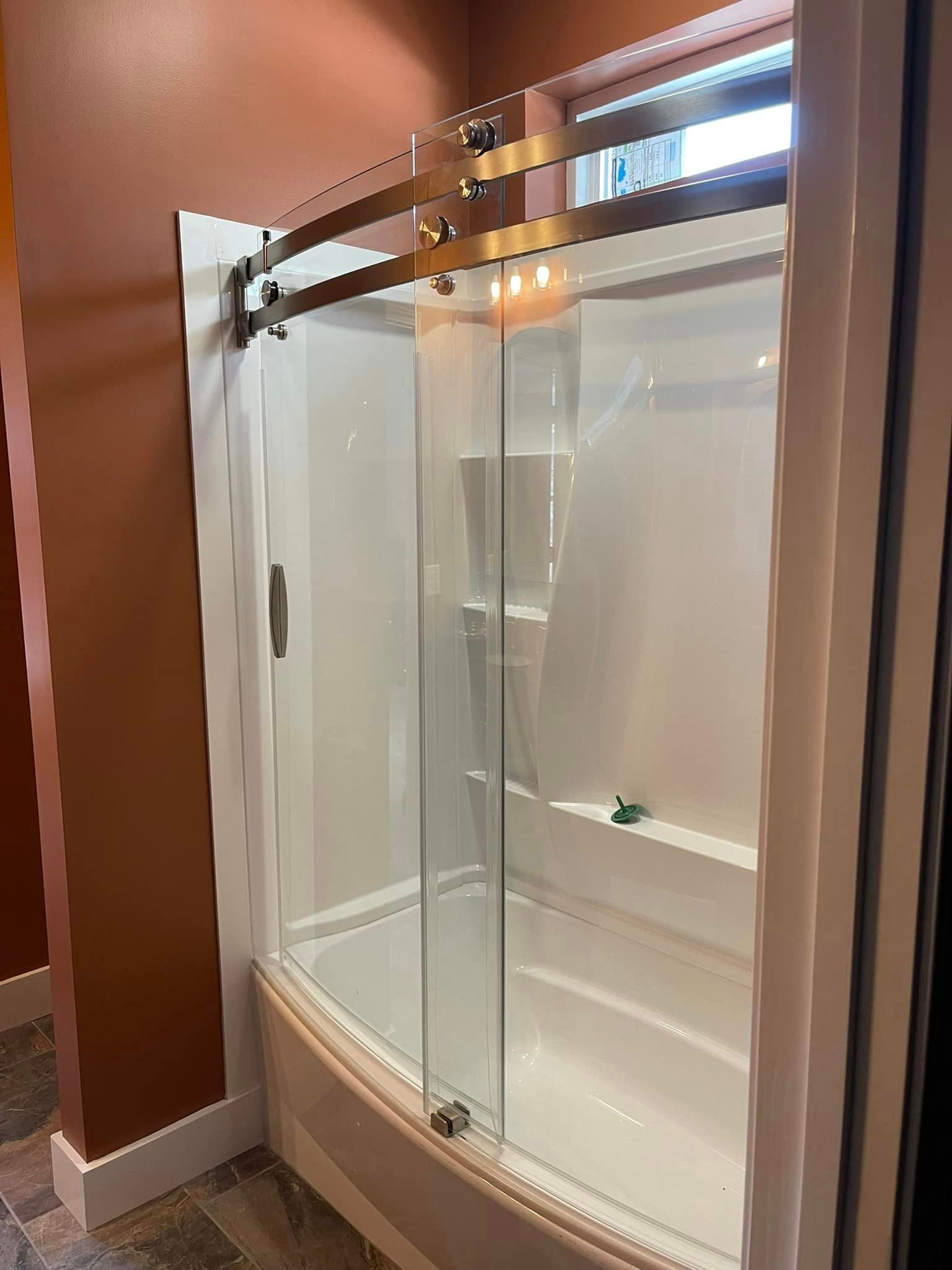 A white bathtub with a glass shower door and bronze fixtures, set in a bathroom with reddish-brown walls.