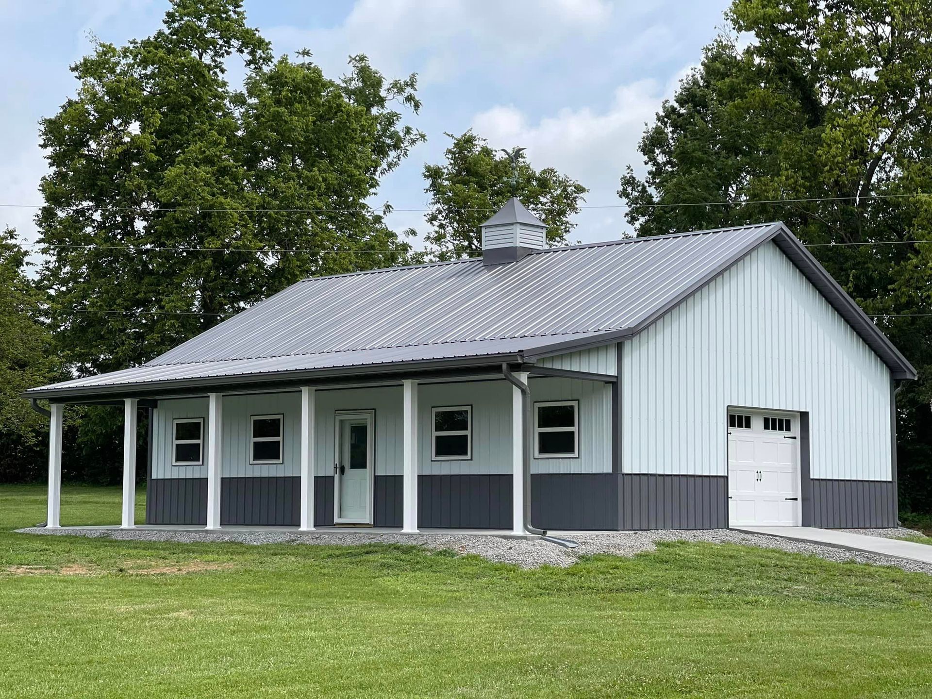 A white and gray metal barn with a porch, a garage door, and a cupola on the roof, set in a grassy field.