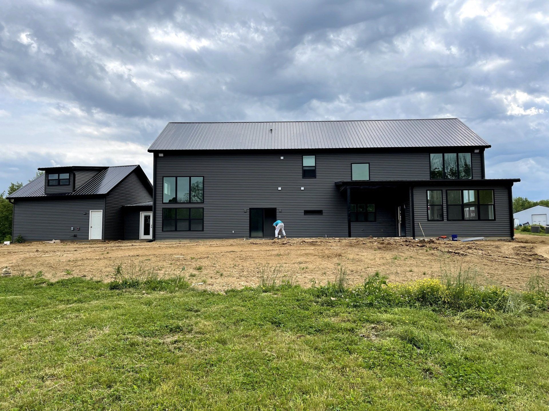 Modern gray house with detached garage under a cloudy sky. Construction site with bare earth and some grass in foreground.