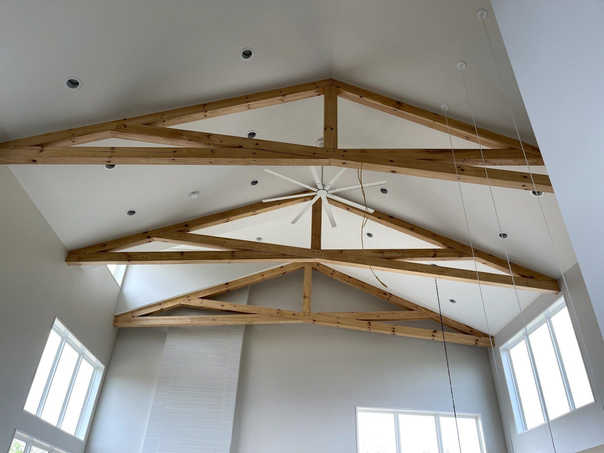 Interior view of a light-filled room with three large wooden trusses on the ceiling. White walls, windows, and a ceiling fan are visible.