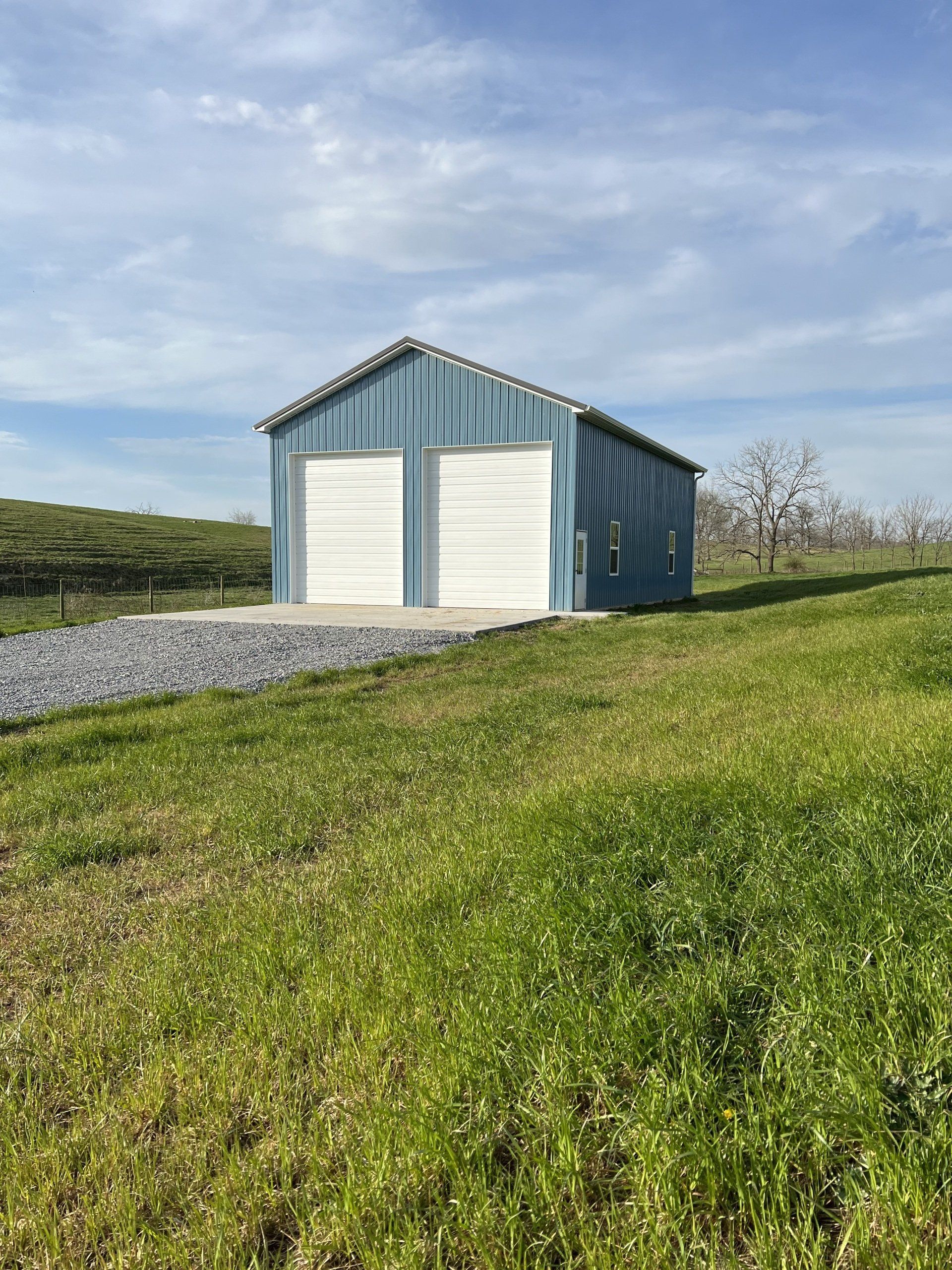 Blue metal shed with two white garage doors on a grassy hill under a partly cloudy sky.